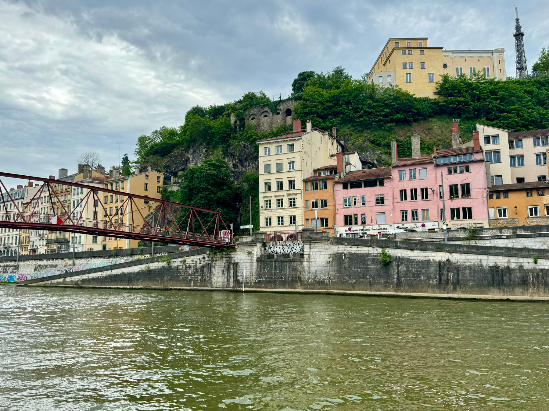 A river in the foreground with a pedestrian bridge to the left. Colorful buildings line the riverbank, backed by a hill with greenery and a large building on top. A tower is visible behind the hill.
