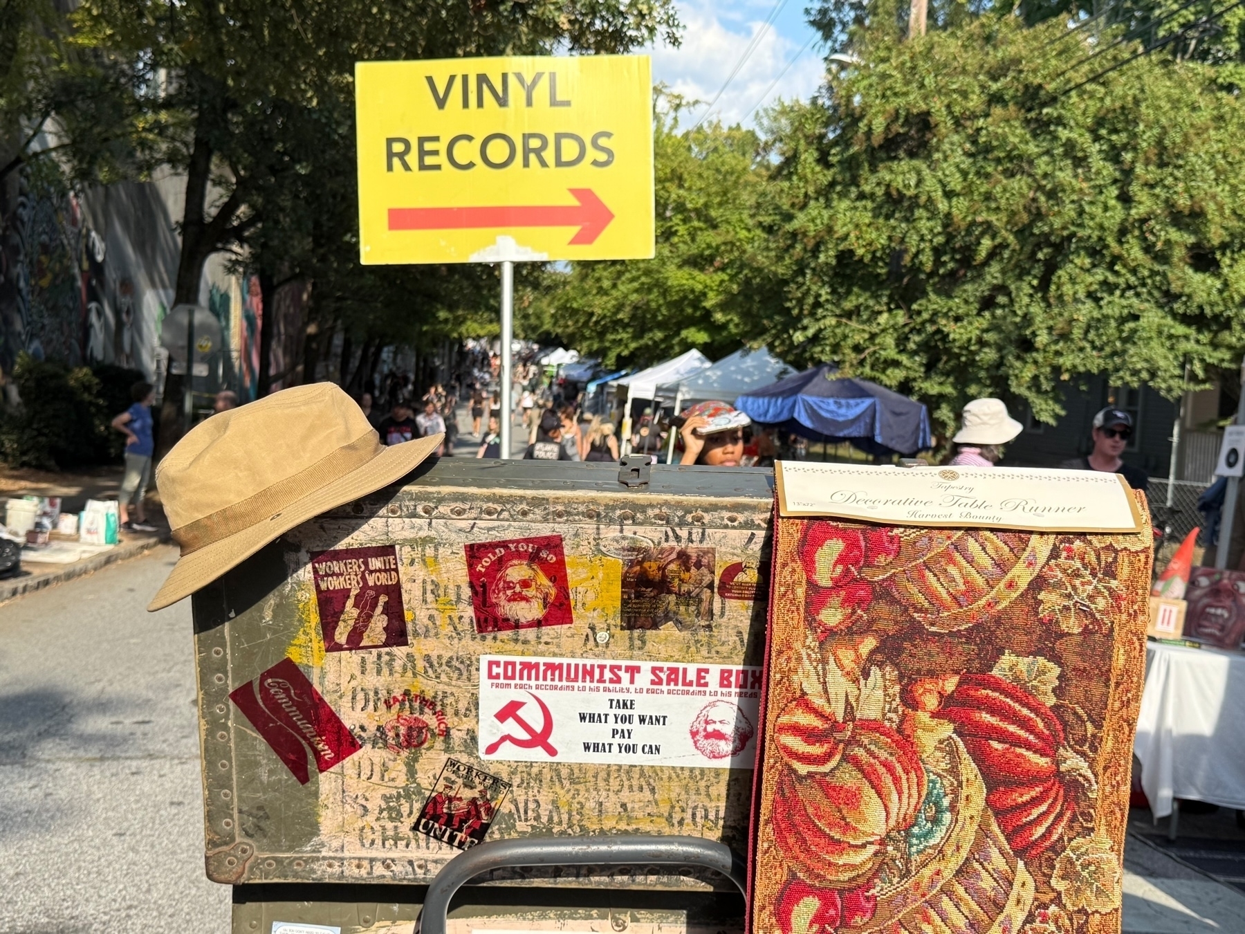 A street market scene featuring a sign pointing to vinyl records. In the foreground, there’s an old suitcase decorated with various stickers, including one that says “COMMUNIST SALE BOX” with the hammer and sickle symbol. A beige hat rests on the box’s corner. 