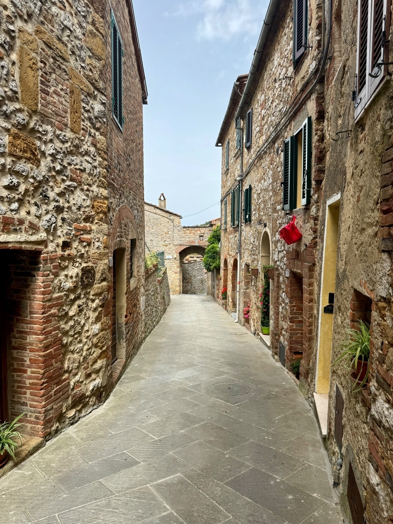 A narrow cobblestone alleyway lined with rustic stone buildings featuring green shutters and flowering plants. The alleyway curves gently with an archway visible in the distance under a partly cloudy sky.