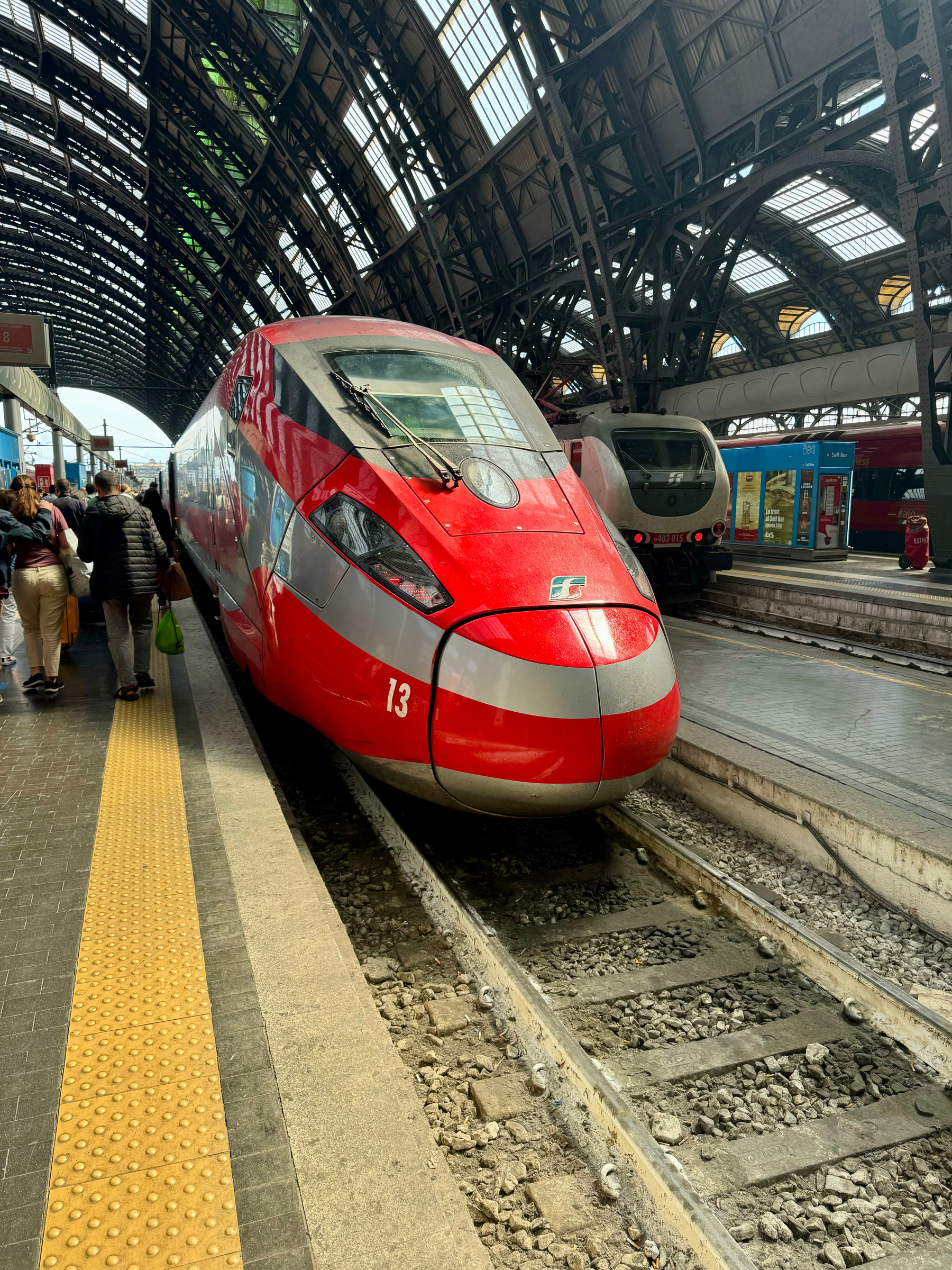 A modern red high-speed train is stationed on a platform in a large, arched train station with a glass and metal roof. Passengers are standing on the platform near the train. A second train is visible on a neighboring track.