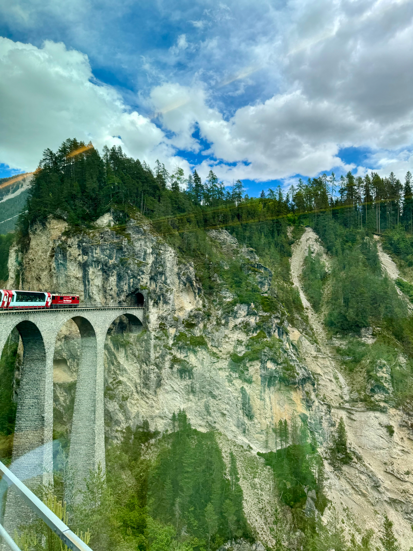 A train crossing a tall, stone arch bridge against a backdrop of steep, rocky slopes and trees under a partly cloudy sky.