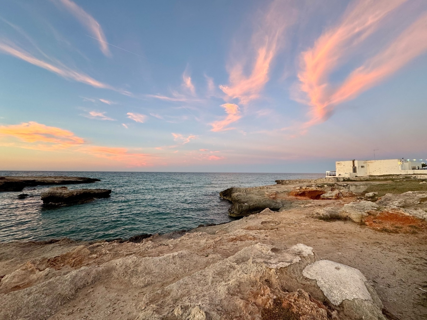 A rocky coastline at sunset with a calm sea. The sky features pastel pink and orange clouds scattered across a blue backdrop. A white building is situated near the edge of the rocky shore on the right side of the image.