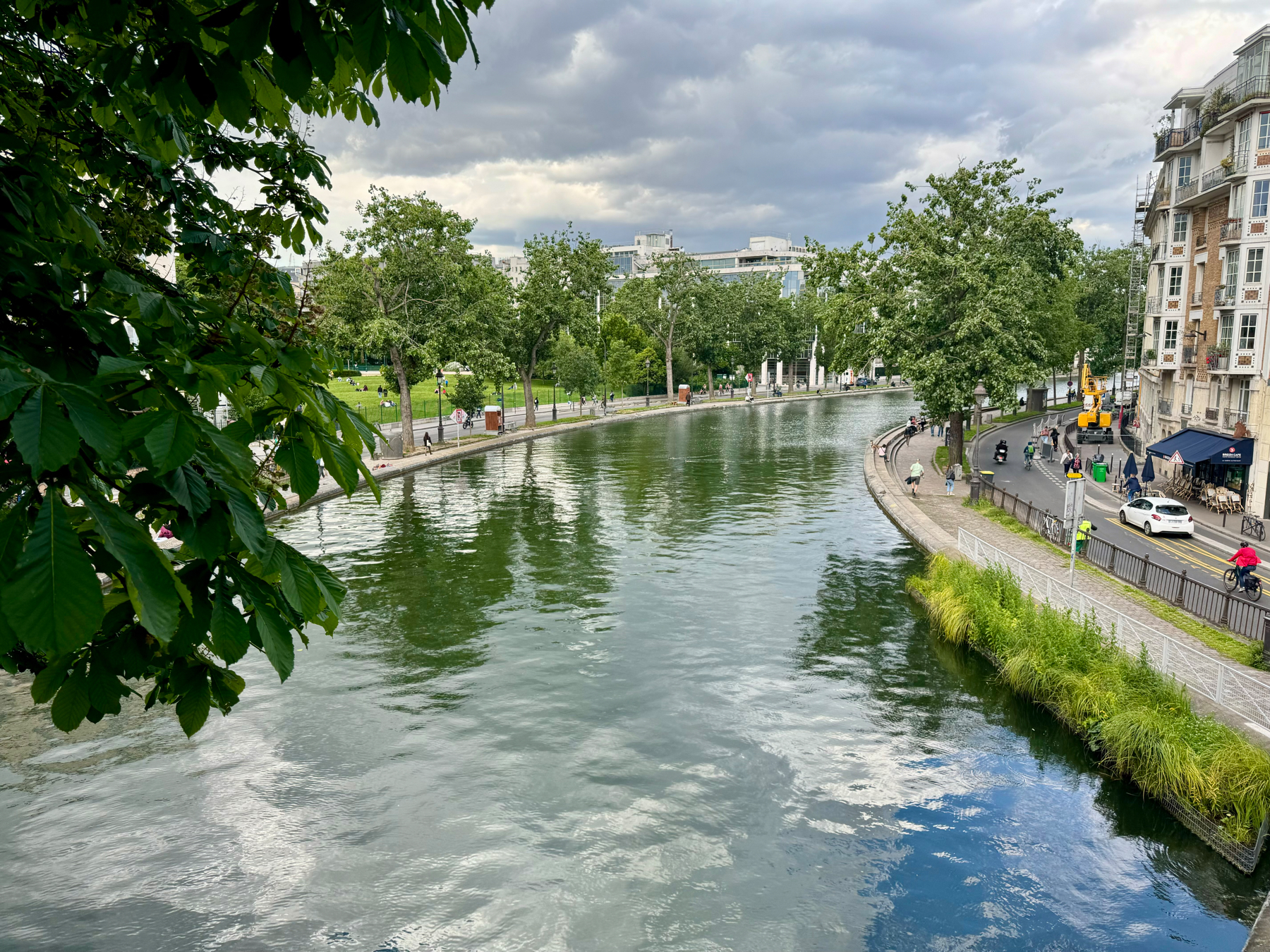 A scenic view of a canal with clear water, bordered by a pedestrian walkway and a road with a few cars and cyclists. Trees and buildings line the opposite bank under a cloudy sky.