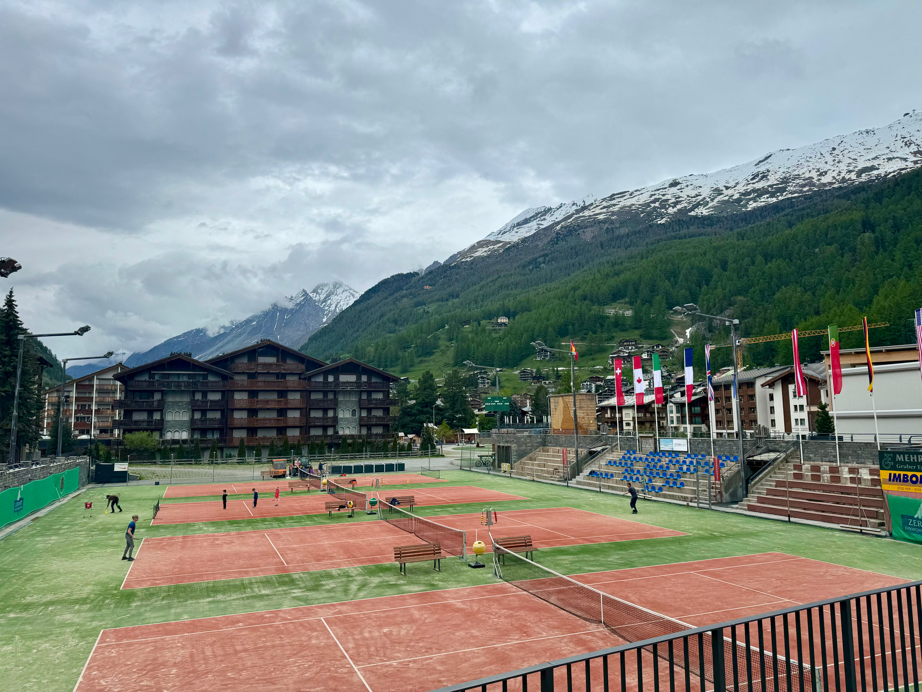 A tennis court in a mountainous region with players on the court, surrounded by buildings and trees, with snow-capped mountains in the background and various international flags displayed.