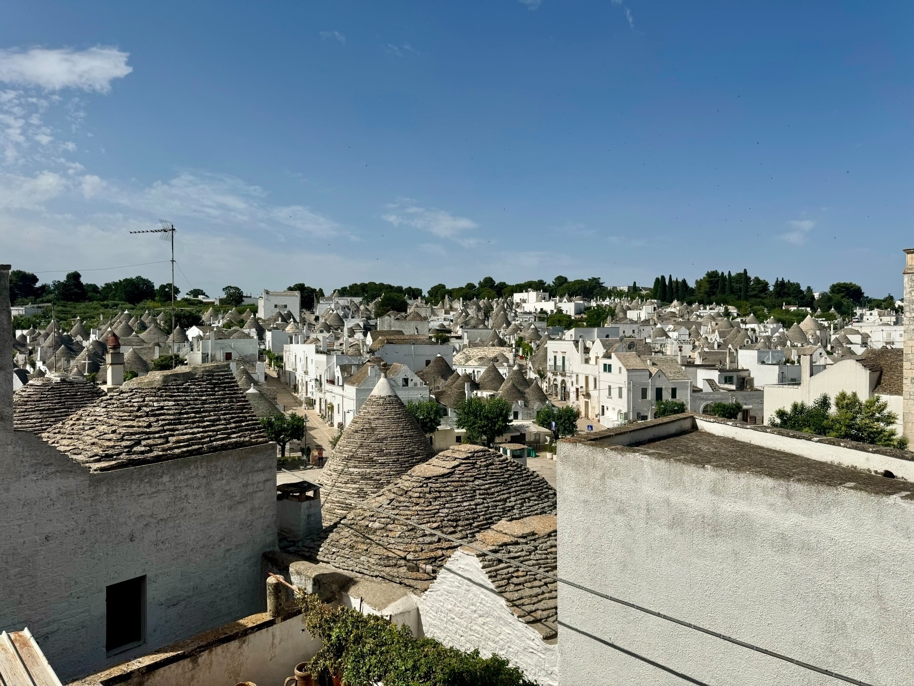 A scenic view of the traditional trulli houses in Alberobello, Italy. The image captures numerous whitewashed stone buildings with distinctive conical roofs under a clear blue sky, with some greenery visible in the background.