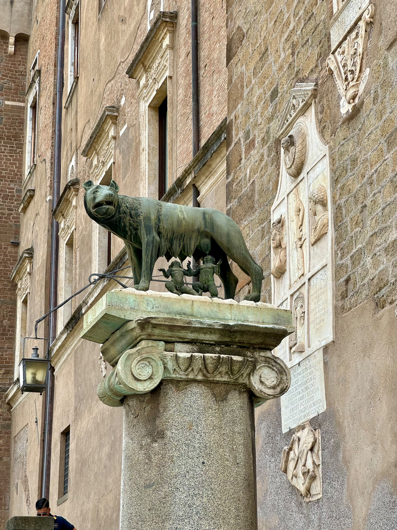 A statue of the Capitoline Wolf suckling Romulus and Remus is mounted on an ornate column in front of an old building with stone and brick walls. The background features sculpted plaques and architectural details. The scene is situated in an outdoor urban setting.