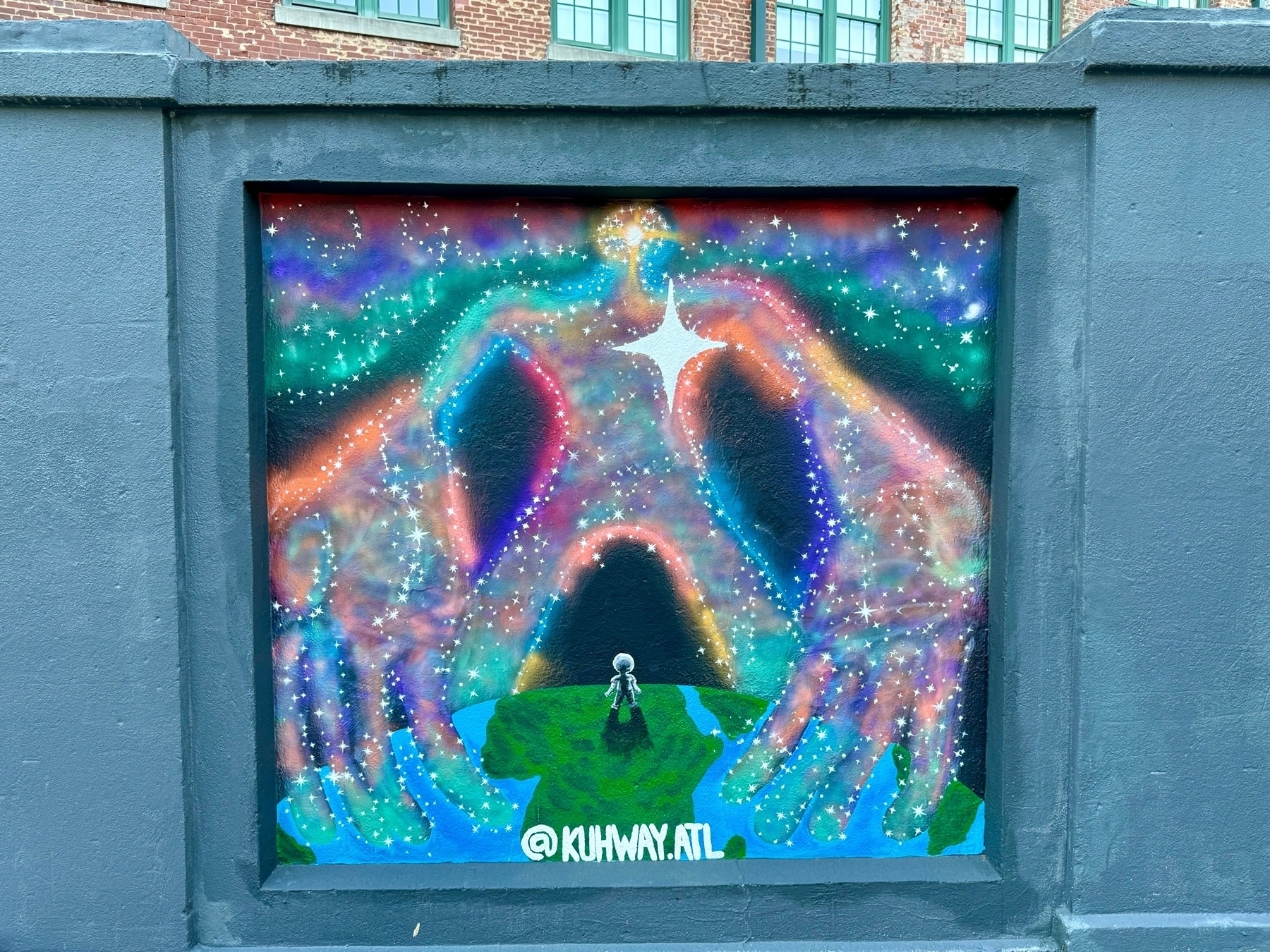 A colorful mural is depicted on a wall, featuring two large, semi-transparent hands crafted from cosmic elements, including stars and nebulous clouds. The hands seem to bend inwards towards a small human figure standing on a green surface, gazing up at it. 