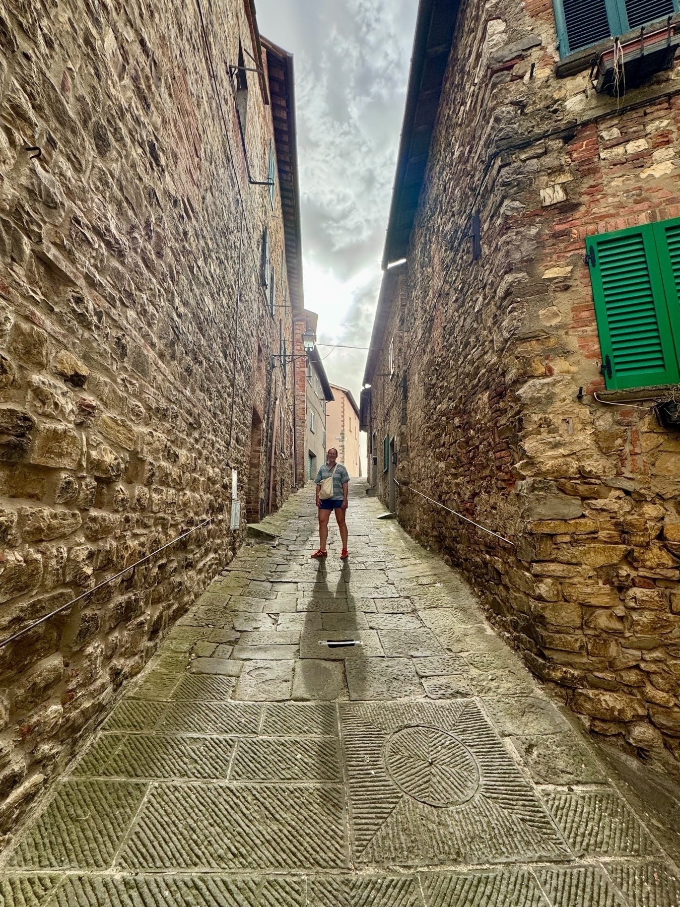 A person stands in the middle of a narrow, cobblestone alleyway flanked by tall, ancient stone buildings with green shutters. The perspective of the alleyway leads the eyes towards a bright, cloudy sky.