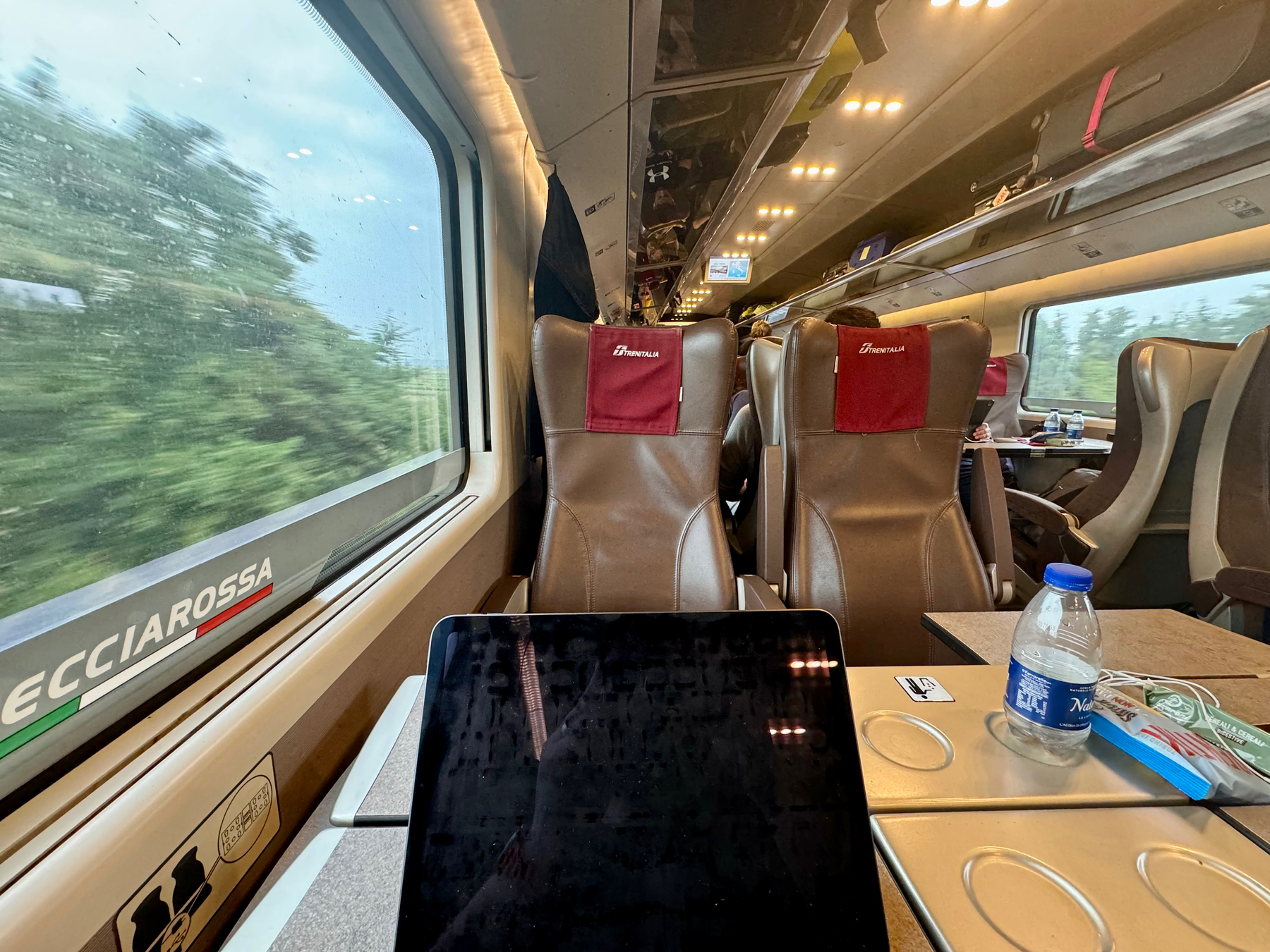 Interior of a high-speed train, specifically a Frecciarossa train by Trenitalia. The image shows rows of seats with red headrest covers, a table with a water bottle, and an electronic device. The train window displays blurred greenery outside. 