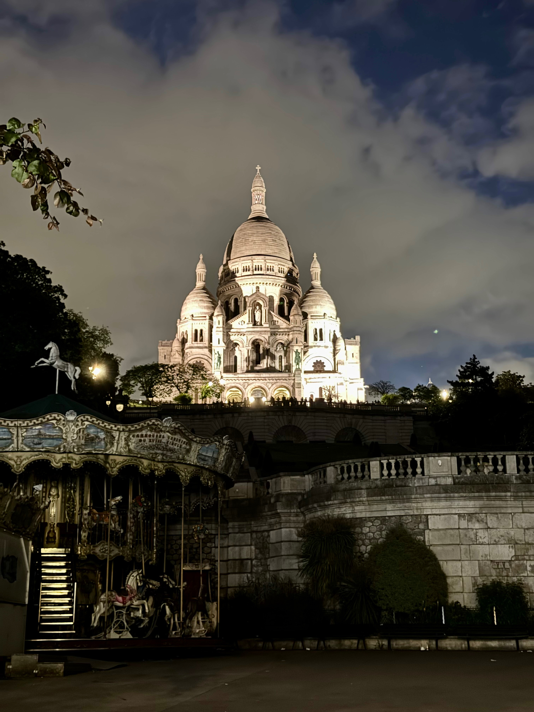The image shows the Basilica of the Sacred Heart of Paris (Sacré-Cœur) illuminated at night, with a vintage carousel in the foreground, set against a backdrop of a dark evening sky with scattered clouds.
