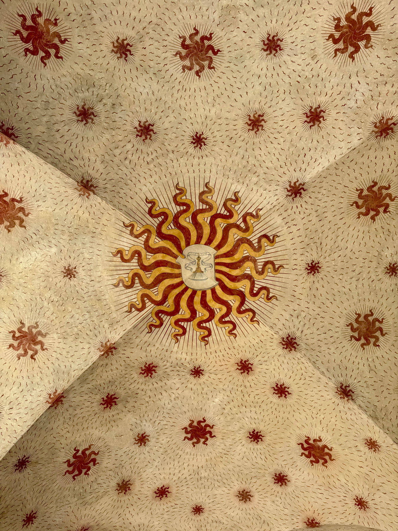 An ornate ceiling design featuring a central sunburst motif with red and yellow rays, surrounded by smaller sunburst patterns. The background is filled with tiny swirling lines, creating a sense of dynamic movement.
