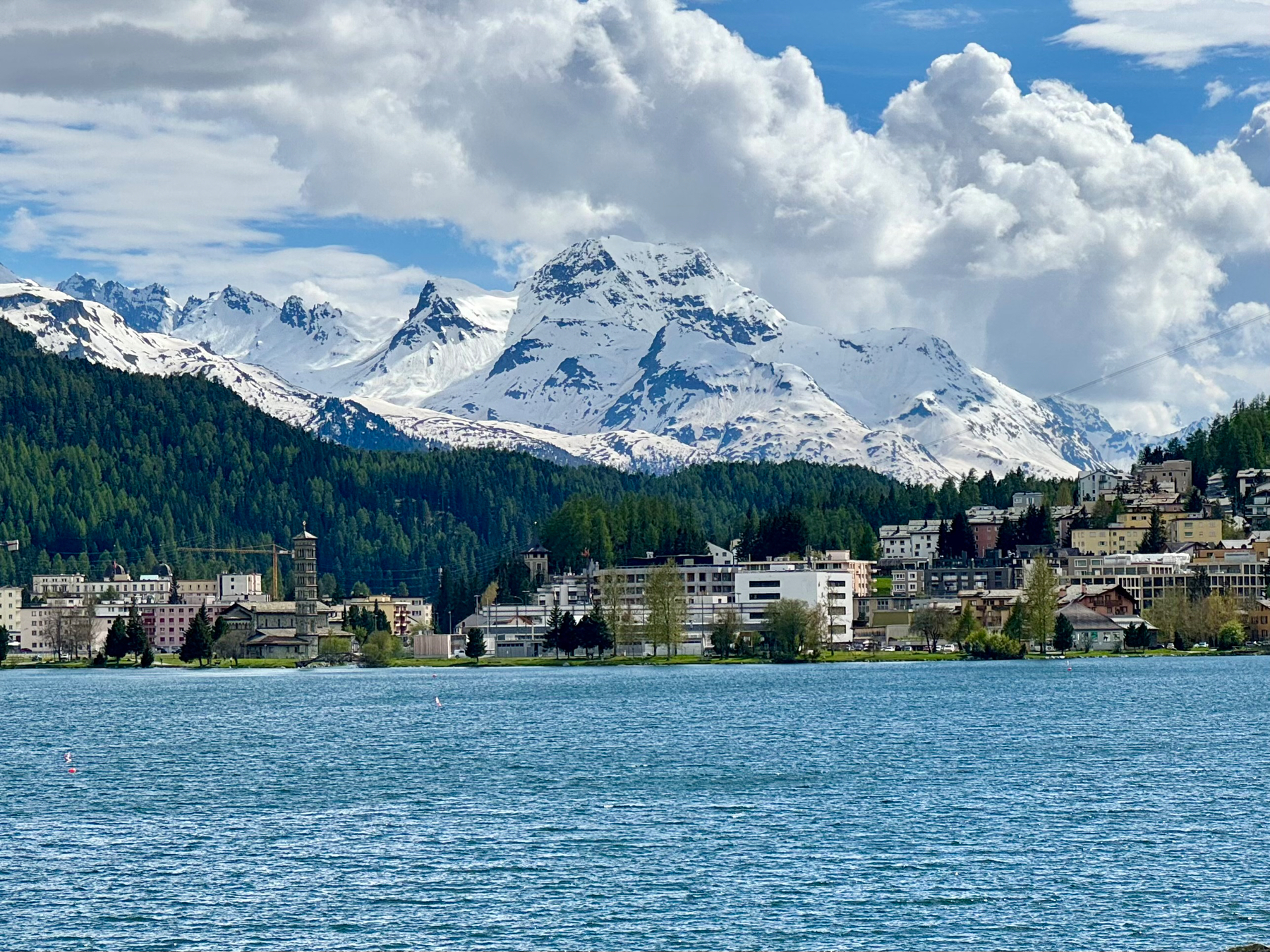 A scenic view of a lake with a town in the foreground and snow-capped mountains in the background under a blue sky with clouds.