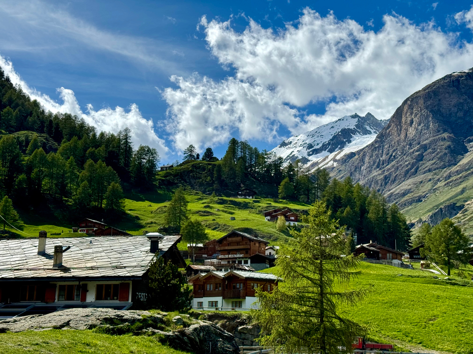 Alpine village with traditional wooden chalets, green pastures, forested slopes, and a snow-capped mountain under a blue sky scattered with clouds.