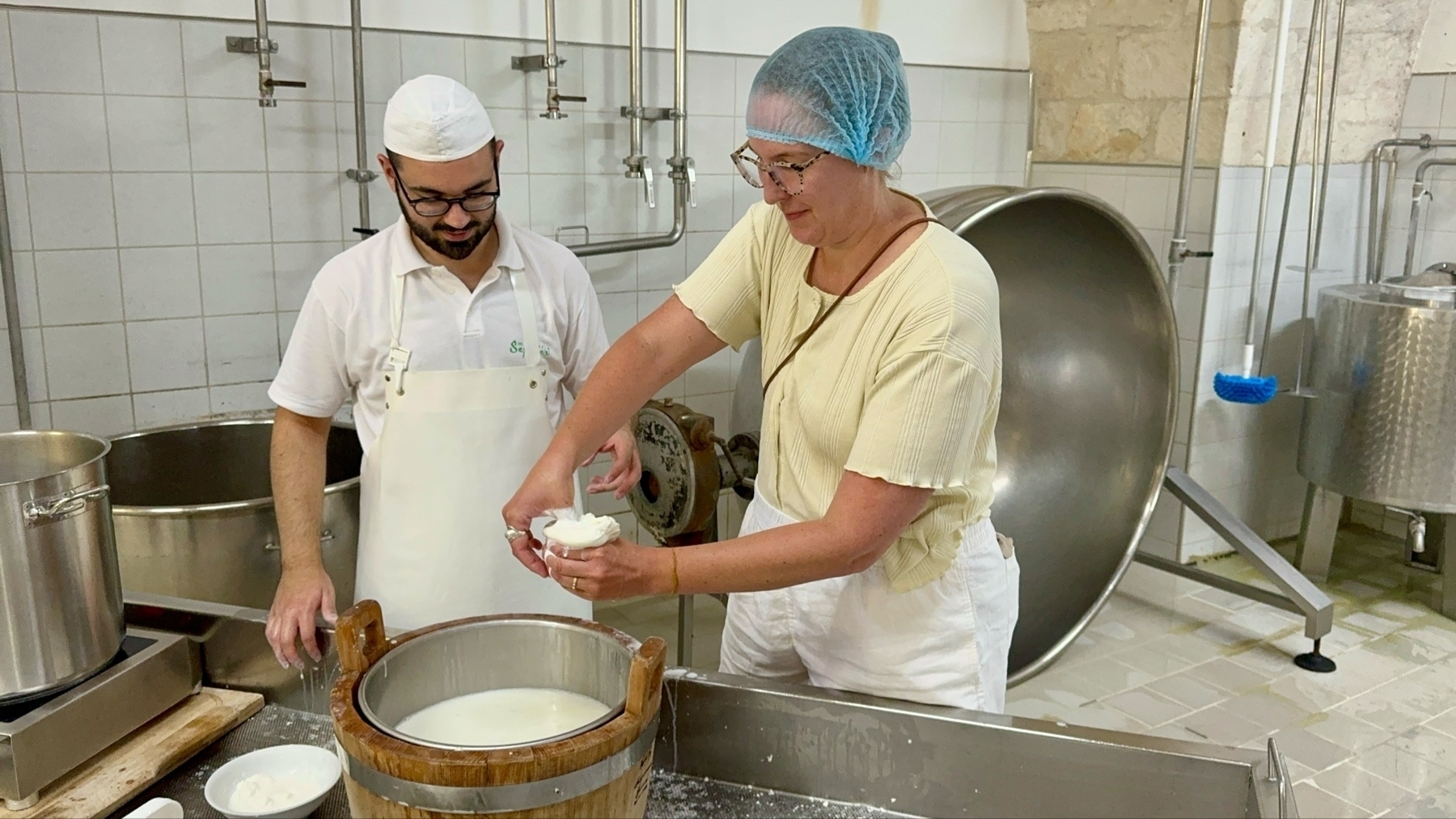 Two individuals are engaged in a cheesemaking process in a dairy facility. They are wearing hygienic hairnets and aprons. One person is pressing curd into a mozzarella ball over a wooden vat filled with milk, while the other observes.