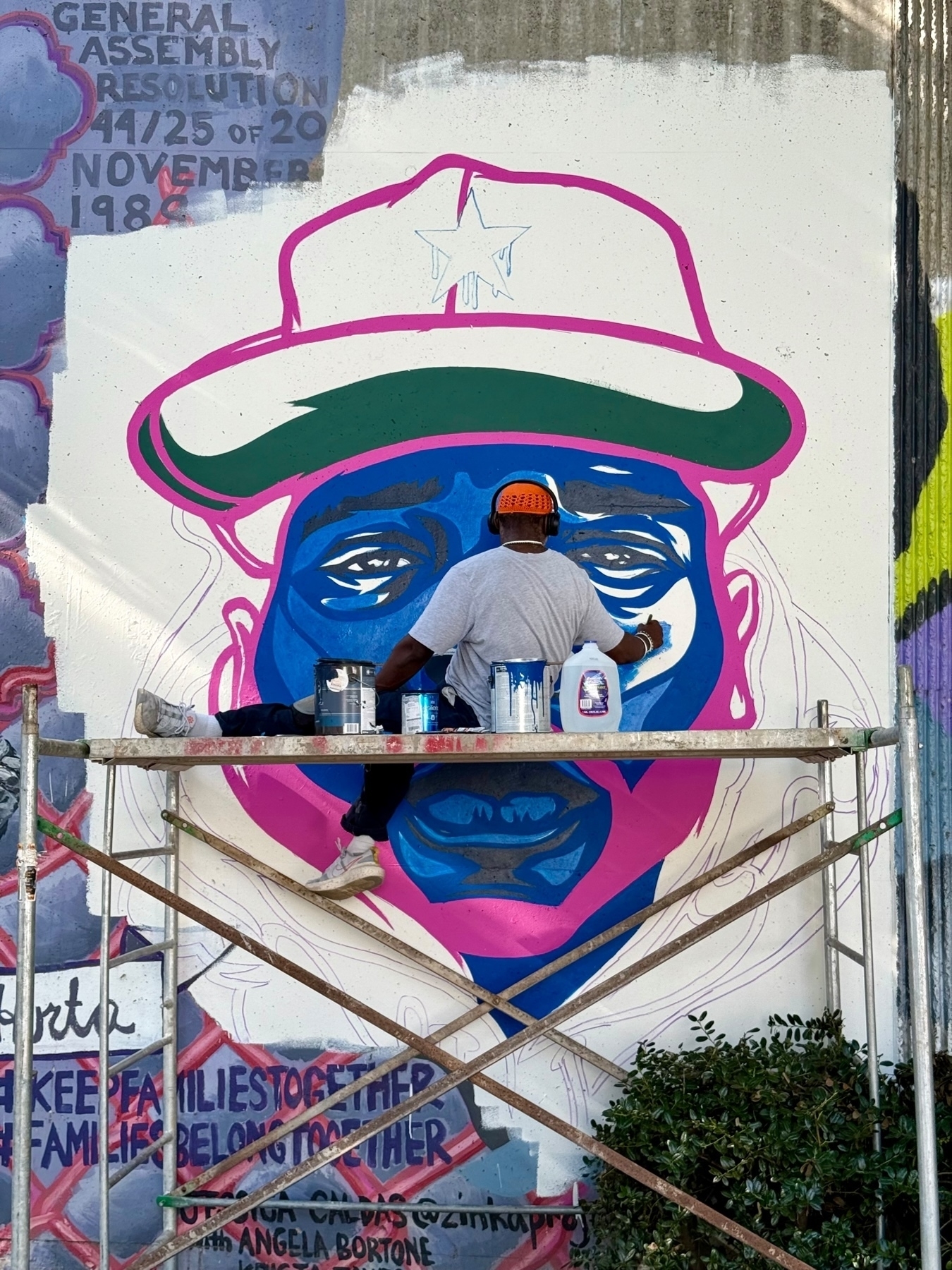 A mural artist is working on a large, colorful portrait featuring a person in a hat with a star on it. The painting is being done on a wall, and the artist is standing on scaffolding with cans of paint and supplies.