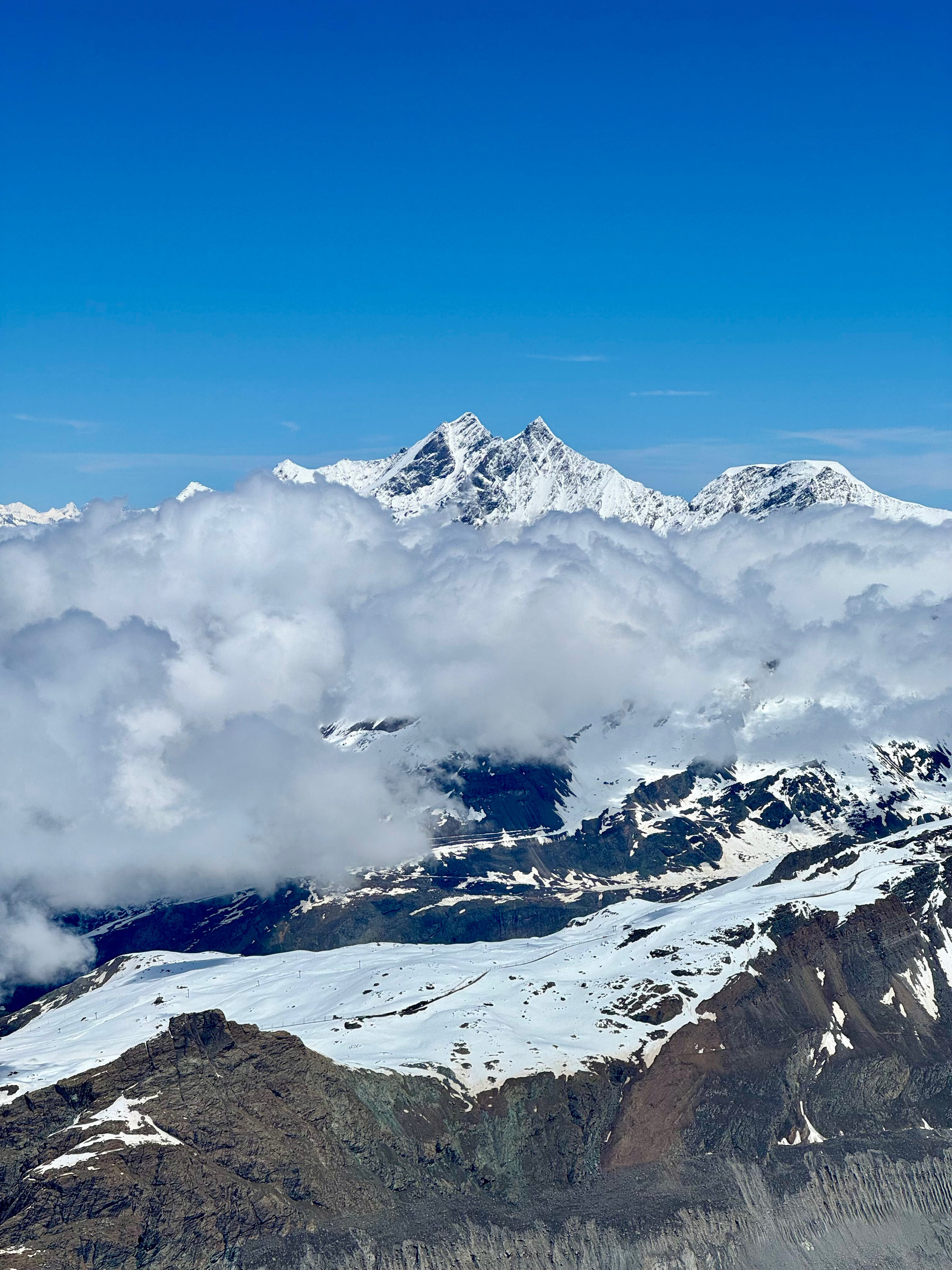 Aerial view of snow-capped mountains piercing through a blanket of clouds under a clear blue sky.