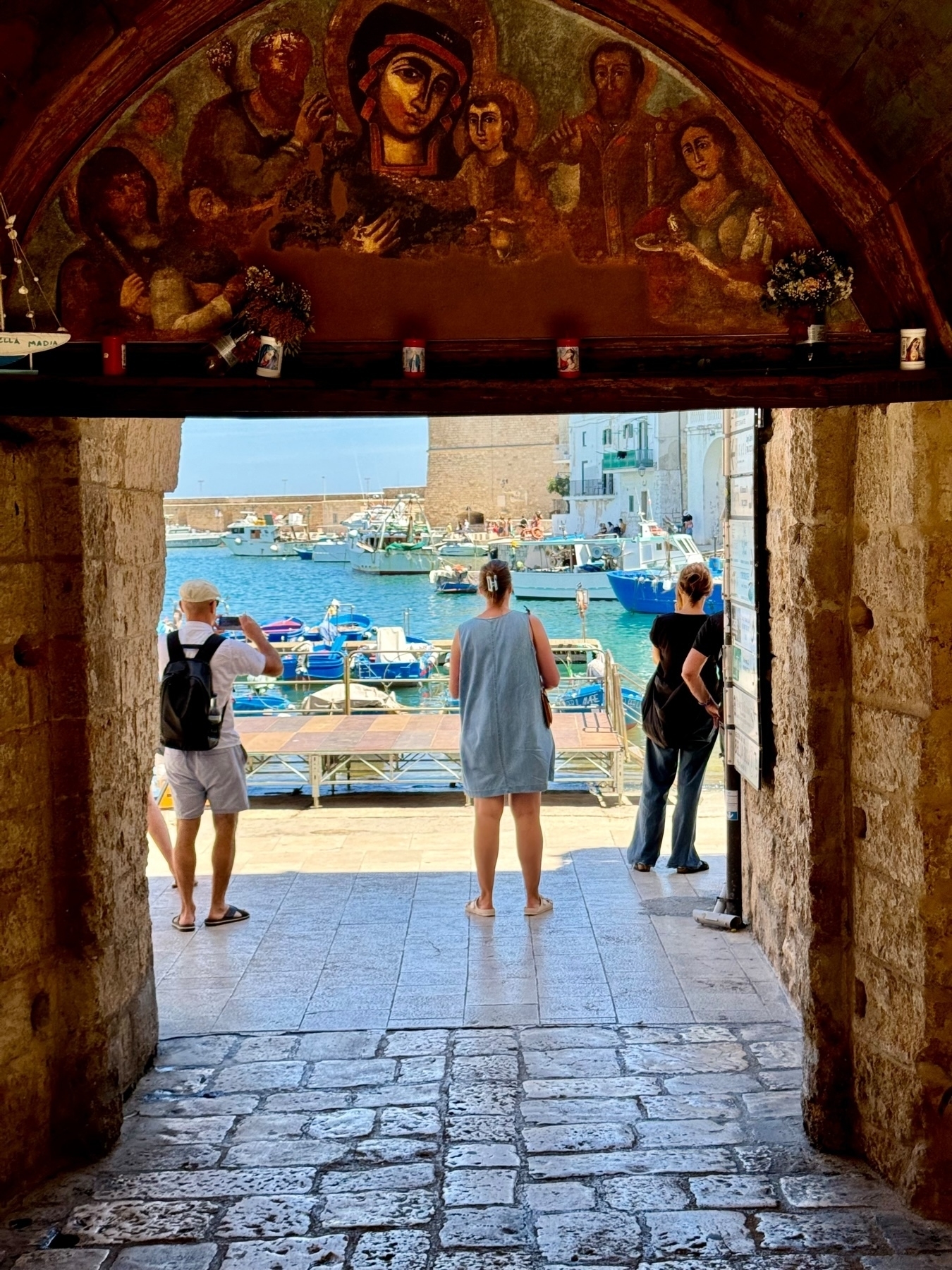 View from a stone archway showing three people looking out at a harbor with boats. Above the arch, there is a mural depicting religious figures. The scene is lively with blue water and boats in the background.