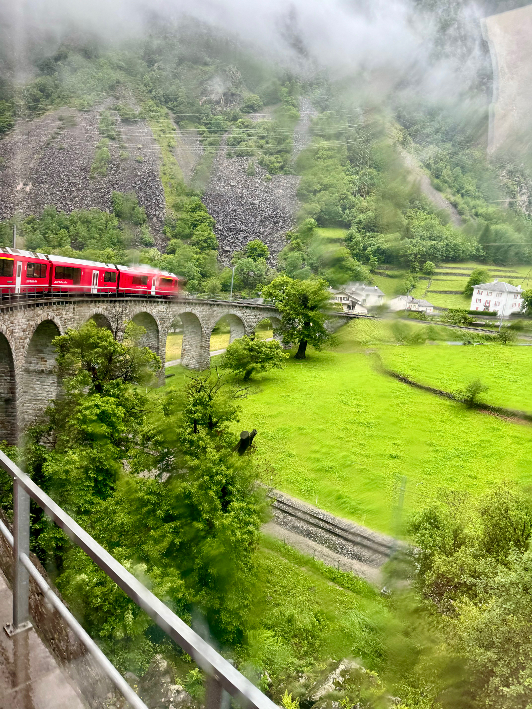 A red train traveling over a stone viaduct surrounded by lush, green landscape and hills. The foreground features a rail, and the image is slightly blurred and streaked, possibly due to raindrops on the camera lens.