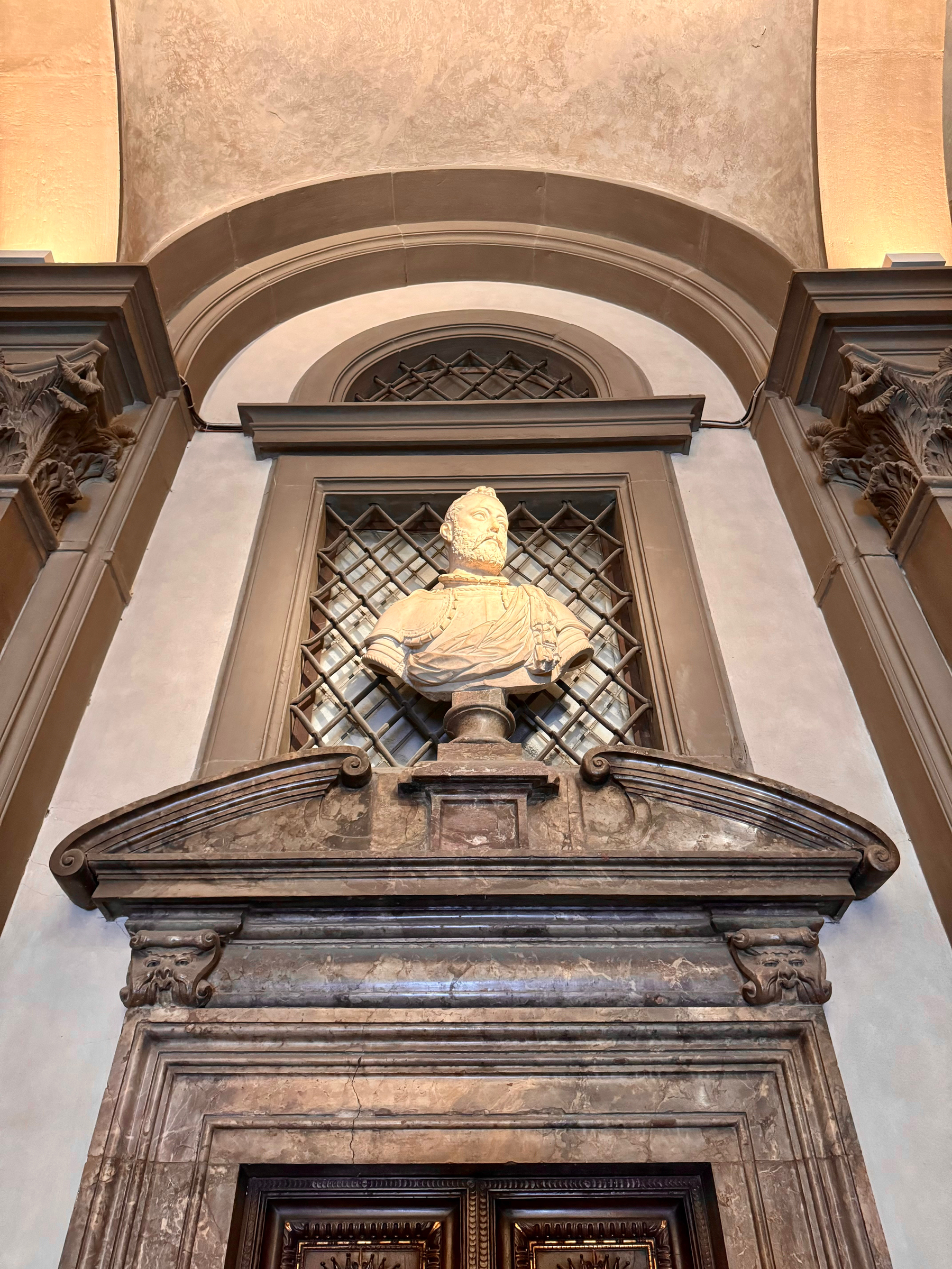 A marble bust of a bearded man in historical attire is placed on a pedestal above an ornately carved wooden doorway. The bust and the door frame are set within an arched architectural niche. The niche has decorative elements including a latticed window. 