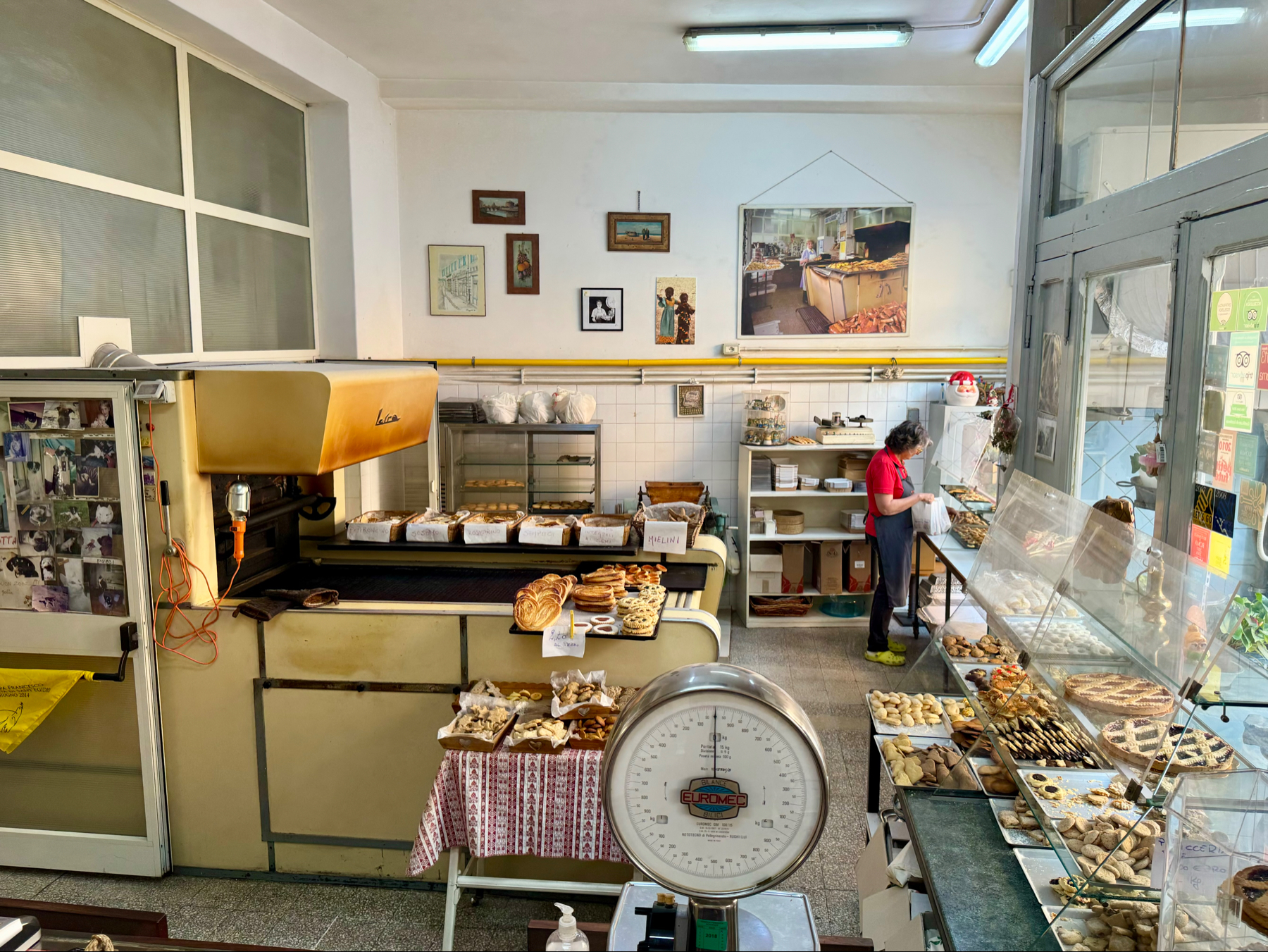 A quaint bakery with a vintage aesthetic. Various baked goods, including bread and pastries, are displayed on counters and shelves. A woman in a red shirt is working behind the counter. The bakery features large baking equipment and a glass display case.