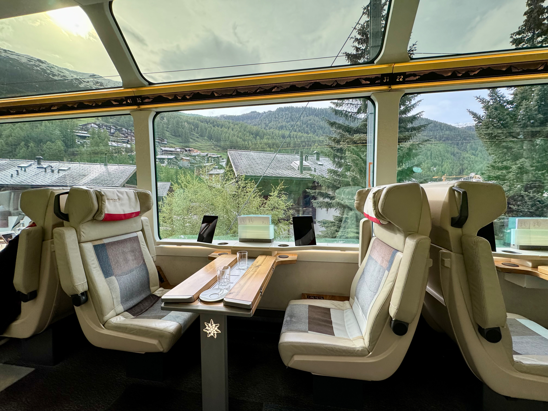 The image shows the interior of the Glacier Express train with spacious seating, large windows providing scenic views of lush greenery, houses, and mountains in the background. The seats are arranged around a central table, with clear glasses placed on top. The train has a luxurious