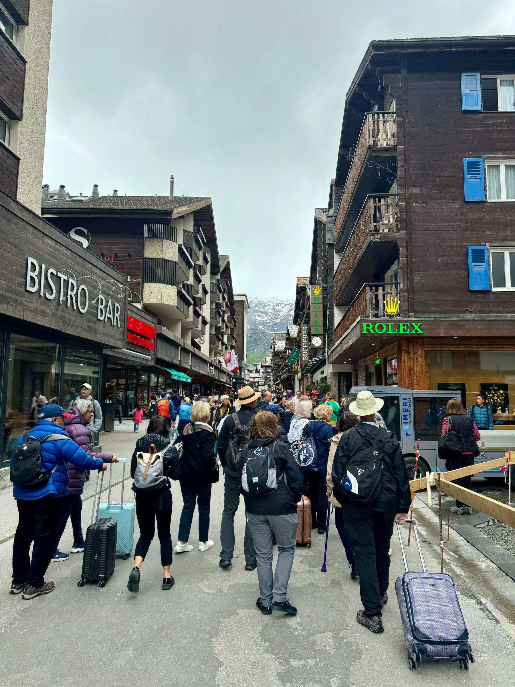 Tourists with luggage walking down a street lined with Swiss chalet-style buildings and shops, including a Rolex store, with mountains in the background.