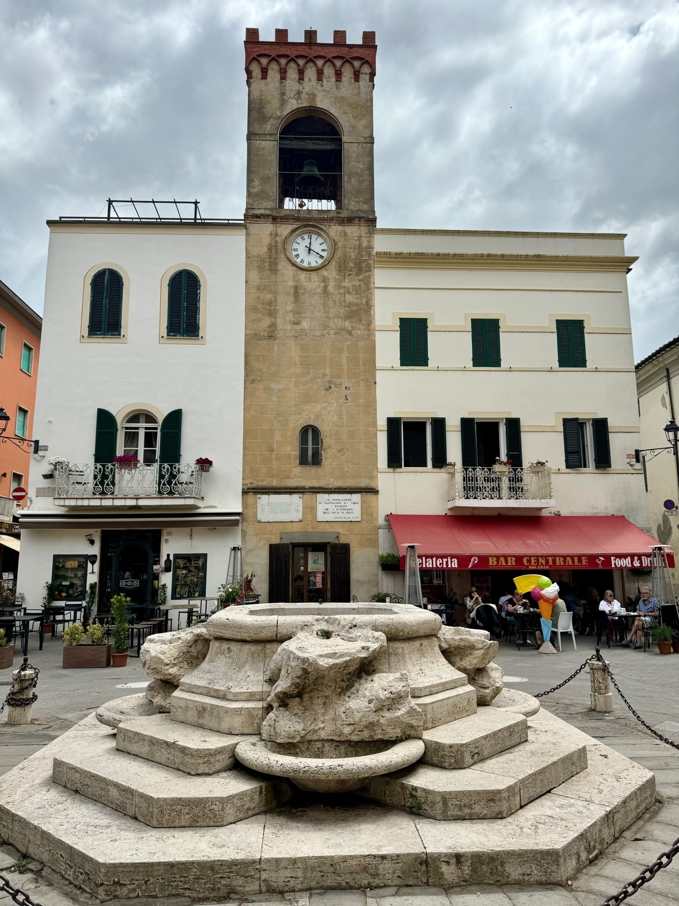 A historical town square featuring a medieval clock tower with a bell, flanked by buildings with green shutters and balconies. In the foreground, there is a stone fountain, and to the right, a café with outdoor seating under a red awning. 