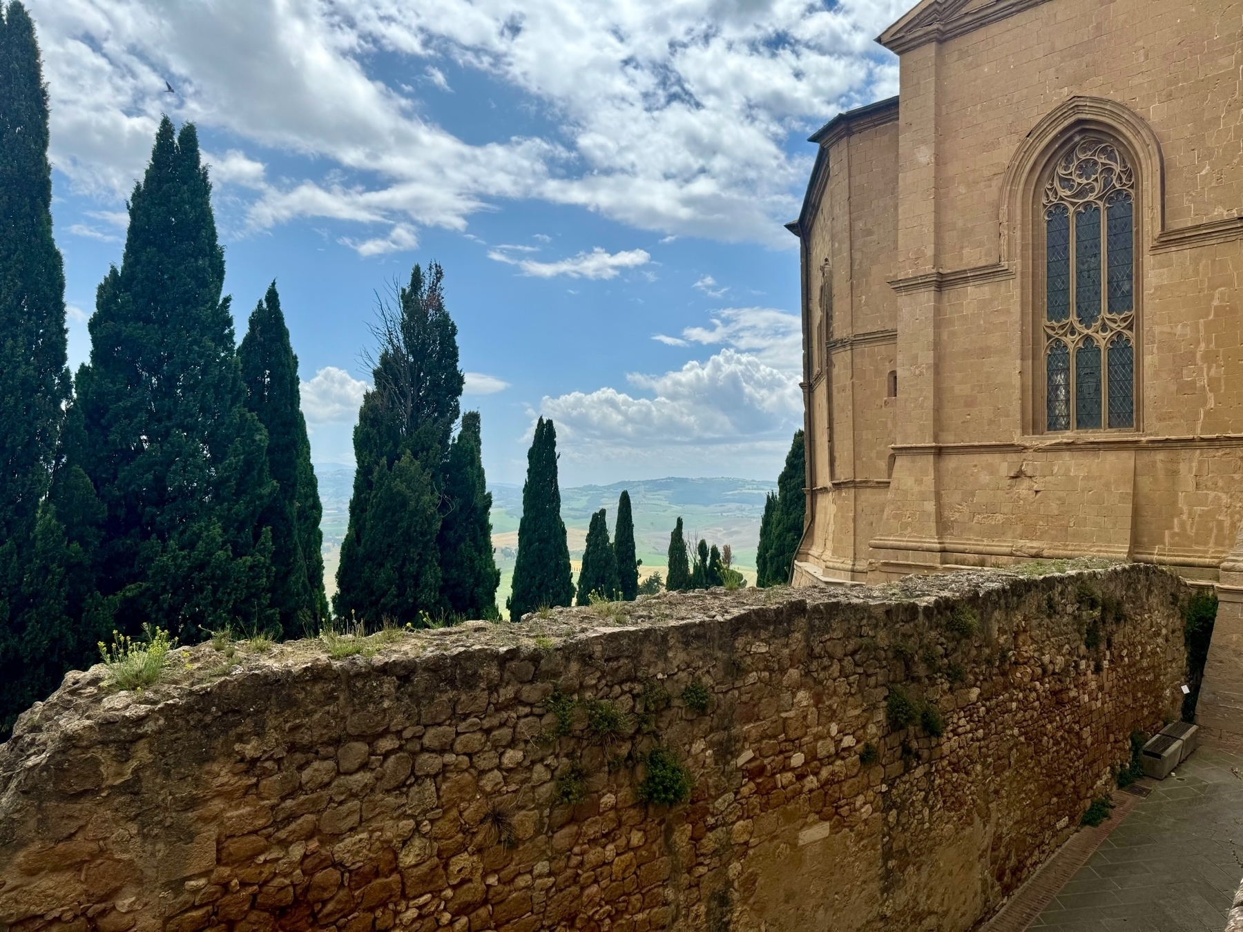 A scenic view featuring a stone wall leading to a large historic building with Gothic-style windows. Tall cypress trees stand beyond the wall, under a blue sky with scattered clouds. The background includes a distant hilly landscape.