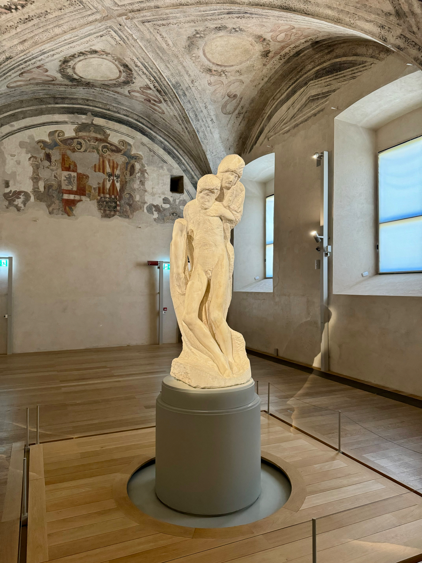 A marble sculpture of two intertwined figures stands in the center of a room with vaulted ceilings adorned with frescoes. The sculpture is displayed on a circular podium with a light wooden floor and has soft natural lighting from two large windows.