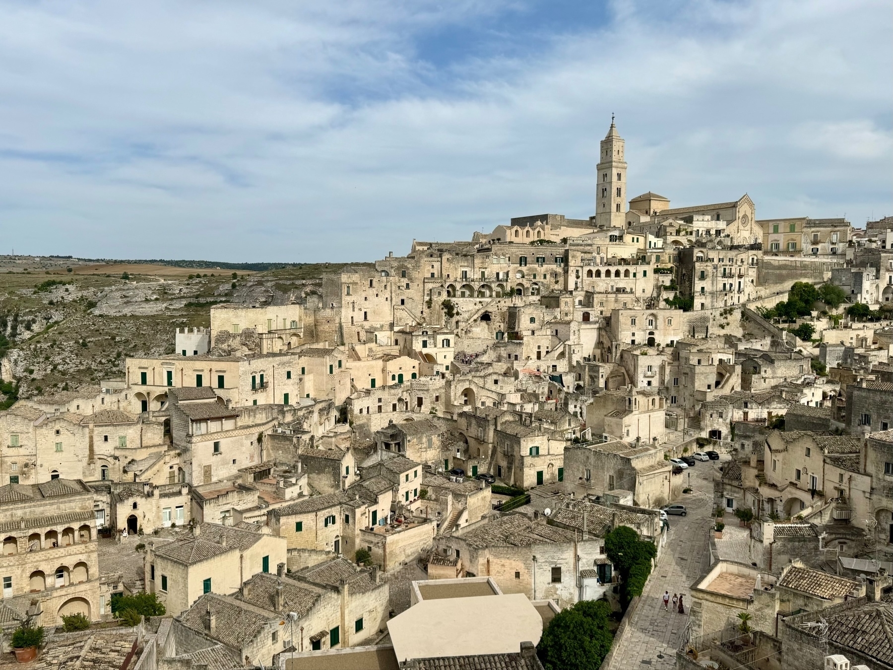 A panoramic view of the historic town of Matera, Italy, featuring ancient stone buildings densely packed on a hillside, winding narrow streets, and a prominent church with a tall bell tower. The landscape around the town consists of rocky terrain and sparse vegetation.