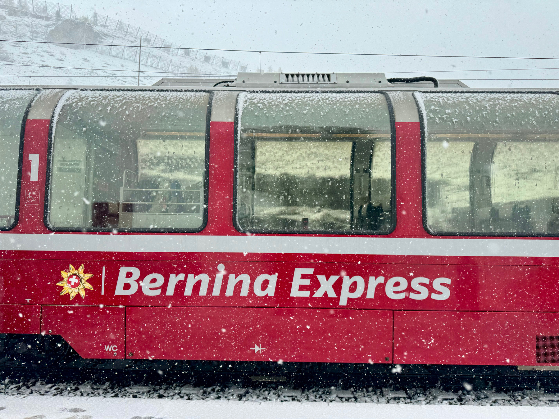 A red, snow-covered train car with large windows, labeled “Bernina Express,” is stopped in a snowy landscape. Snowflakes are visible in the air, and the setting appears to be a cold, wintry environment.