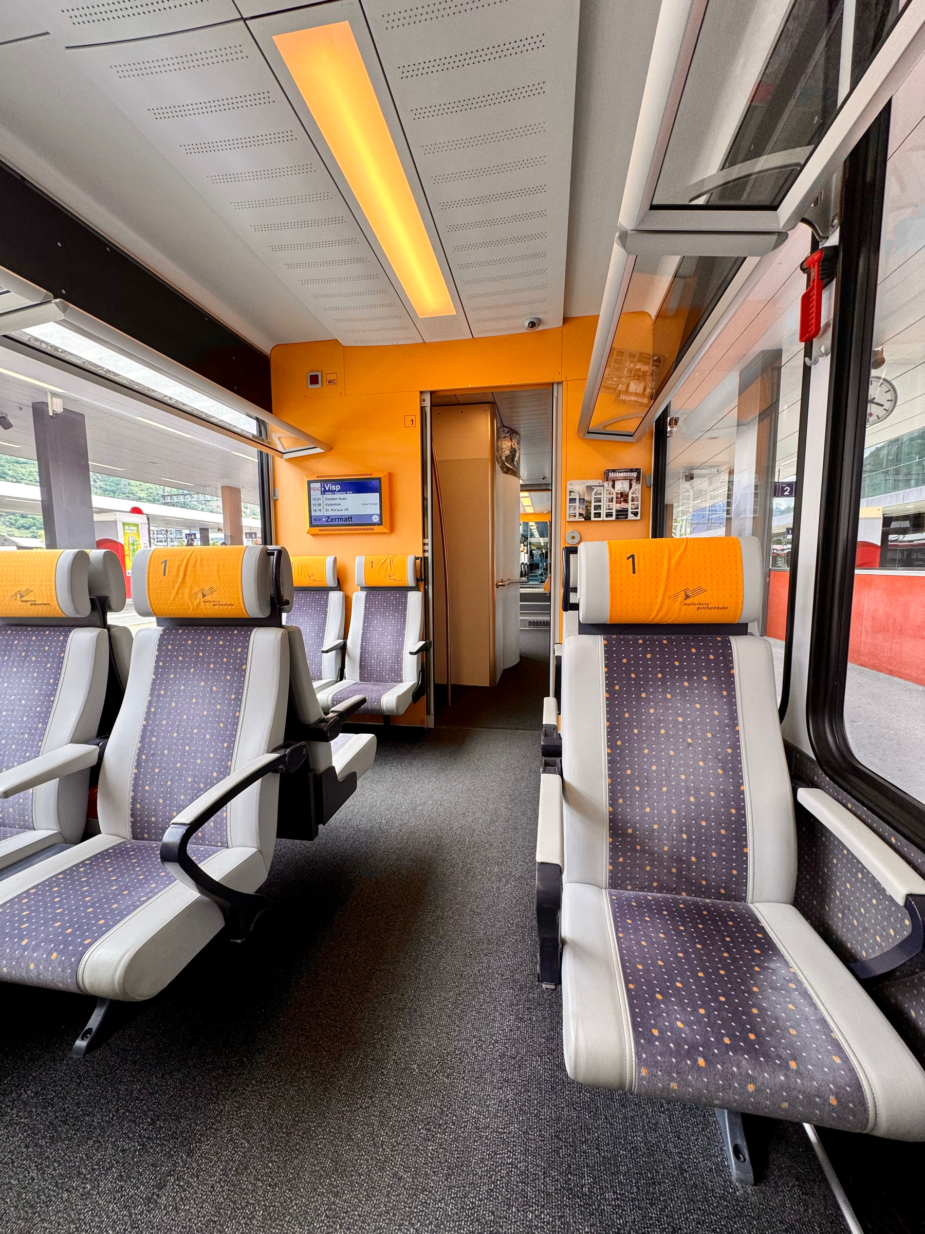 Interior of a modern train carriage with empty seats and large windows, featuring a bright orange and white color scheme.