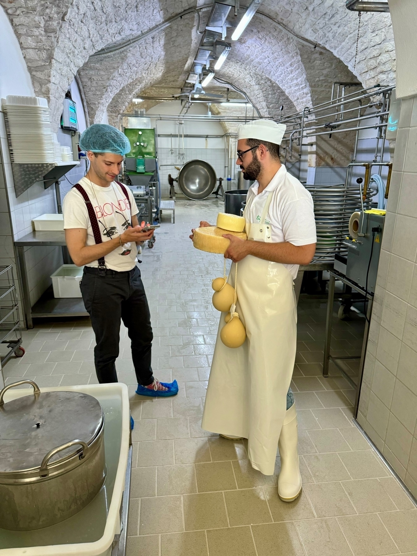 Two people are in a cheese-making facility. One person, wearing a hairnet, suspenders, and shoe covers, looks at their phone. The other person, dressed in white protective clothing, including a hat, apron, and boots, holds cheese. 
