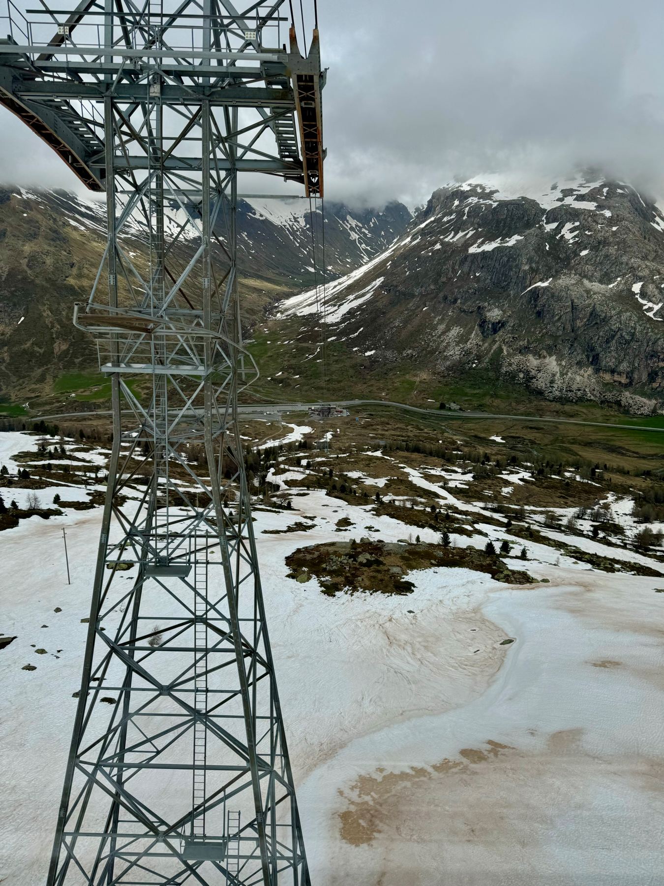 A towering metal cable car structure with a mountainous background partially covered in snow under a cloudy sky.