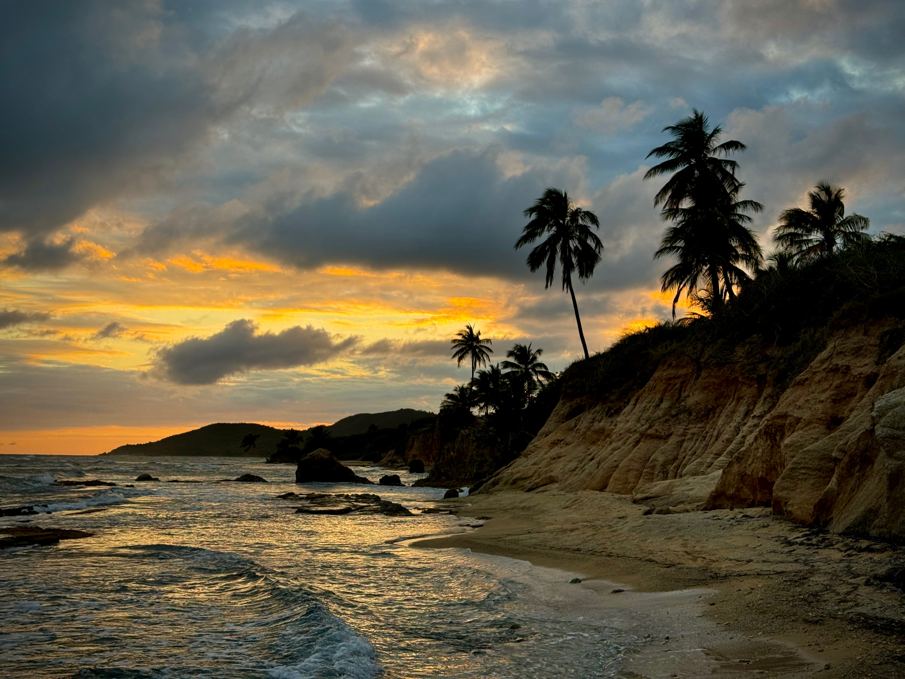 A sunset view over a tropical beach with silhouetted palm trees against a sky with orange-tinged clouds and a calm sea in the foreground.