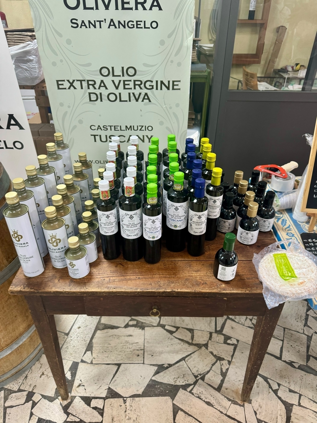 The image shows a display of various bottles of extra virgin olive oil on a small wooden table. The bottles are of different sizes and have different colored caps.