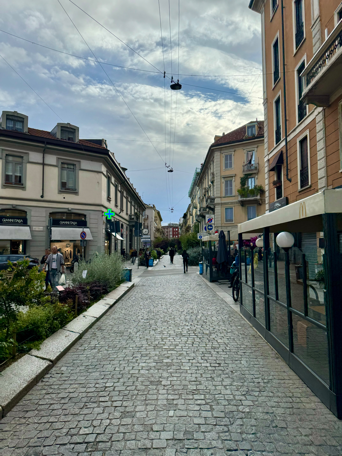 A cobblestone pedestrian street lined with shops and buildings. Overhead, wires cross the partly cloudy sky. Some people are walking and shopping, with greenery and plants adorning the sides of the path.