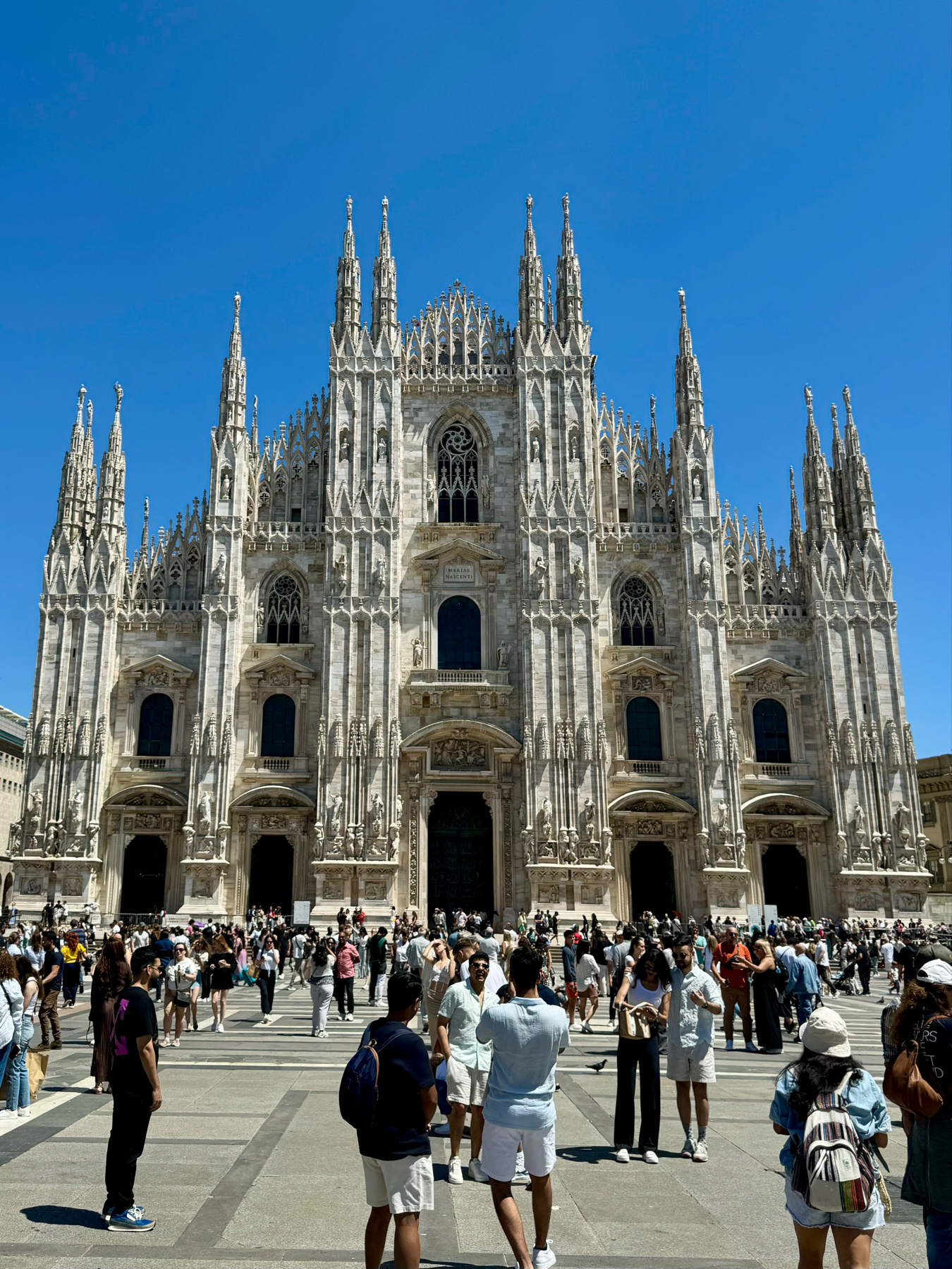 A large crowd of people stands in front of the Milan Cathedral (Duomo di Milano) on a sunny day. The Gothic architectural details of the cathedral are prominent against a clear blue sky. Tourists are visible taking photos and appreciating the historic structure.