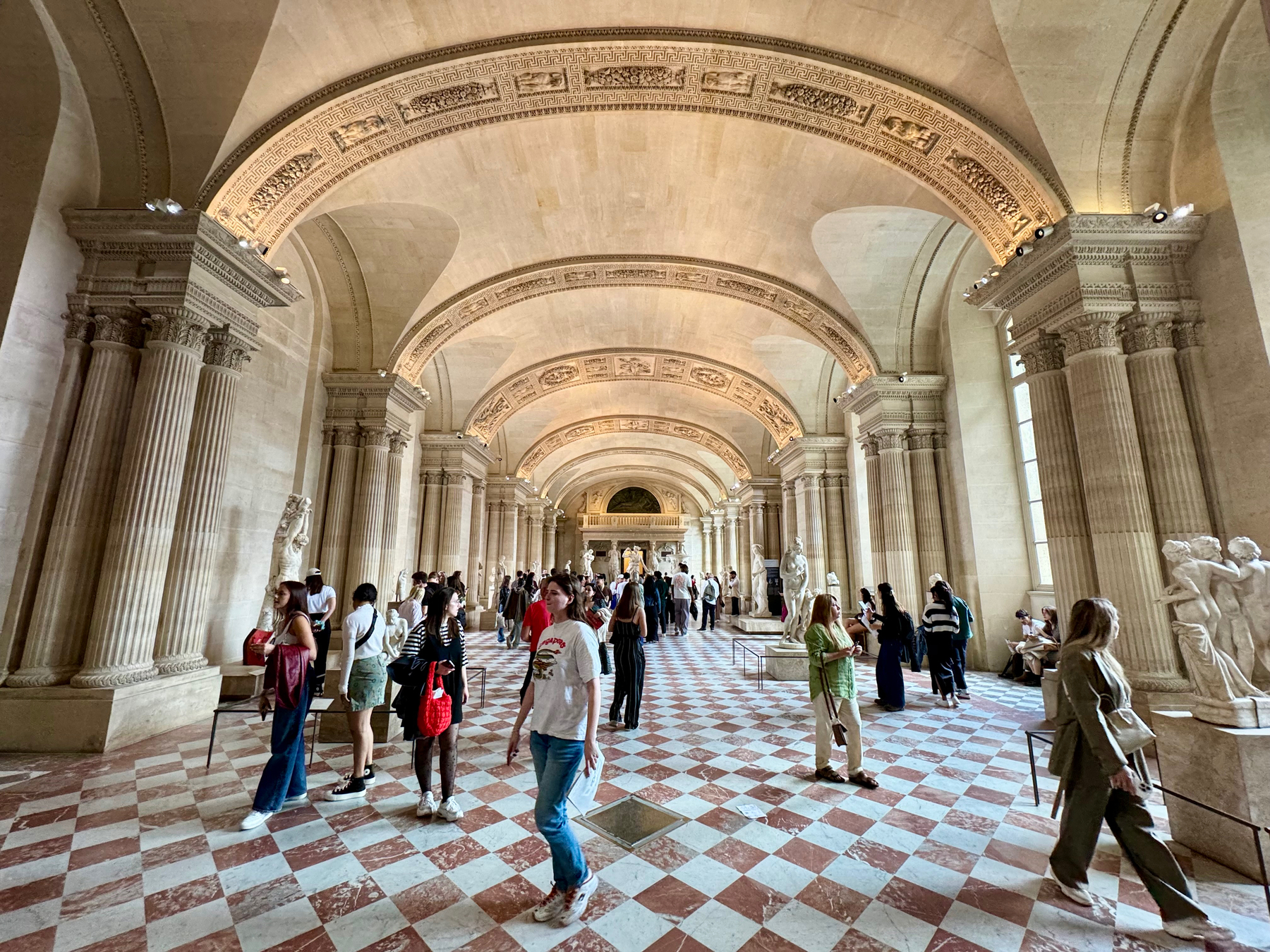 Interior of a museum gallery with classical architecture, featuring columns and sculptures, with visitors walking and observing the art.