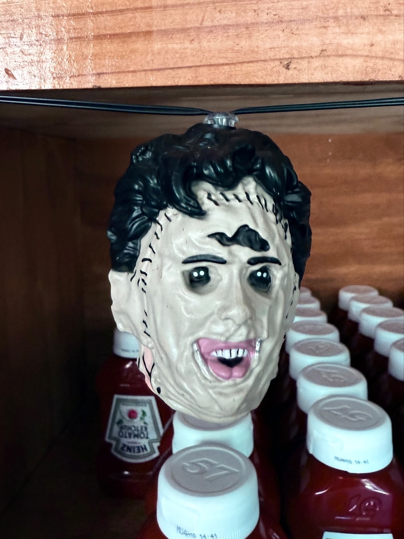 A plastic mask resembling a horror character with stitched facial features is hanging above rows of Heinz ketchup bottles on a wooden shelf.