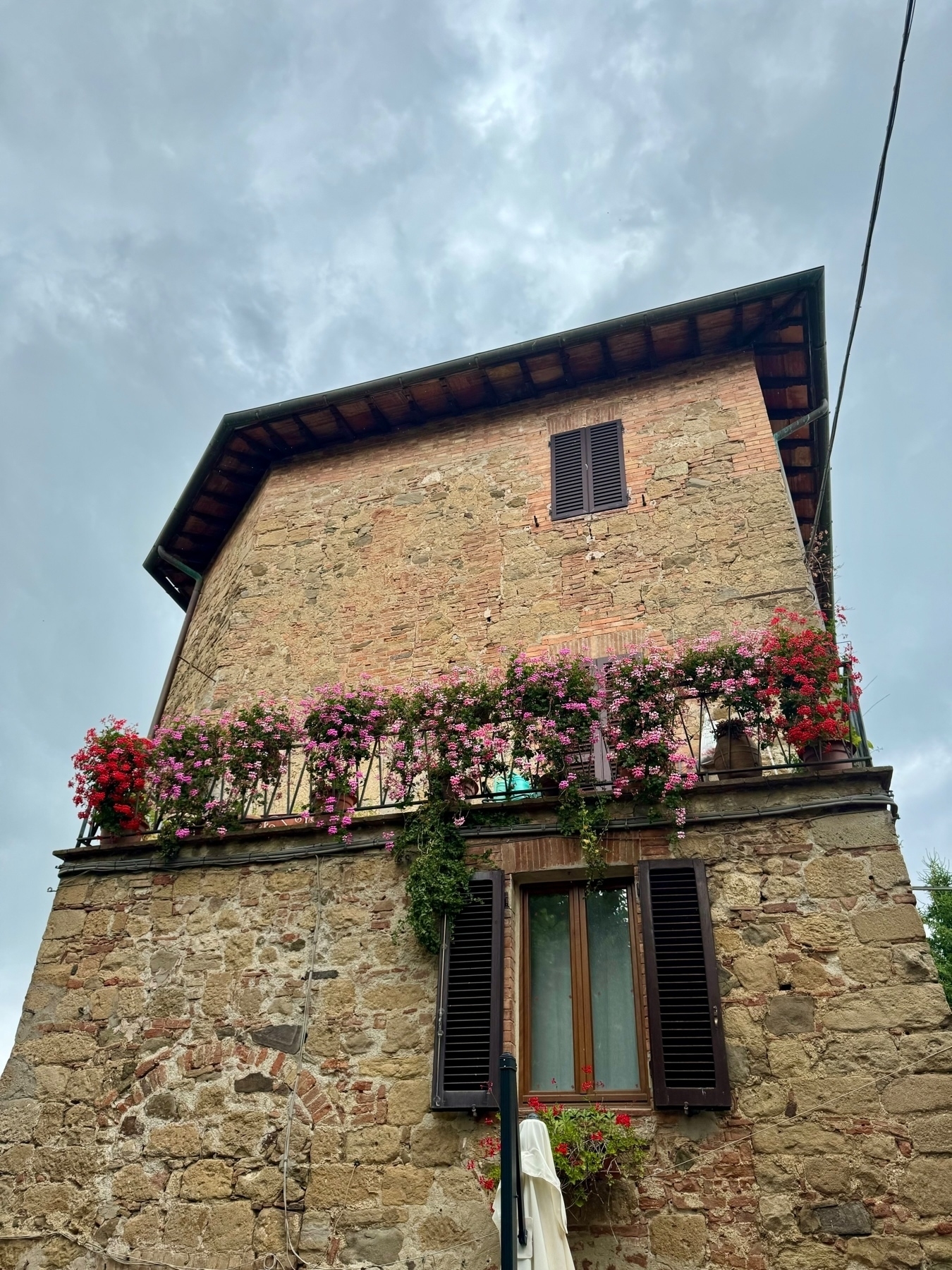 A rustic stone building with wooden window shutters, featuring vibrant flowers hanging from a balcony. The sky is overcast.