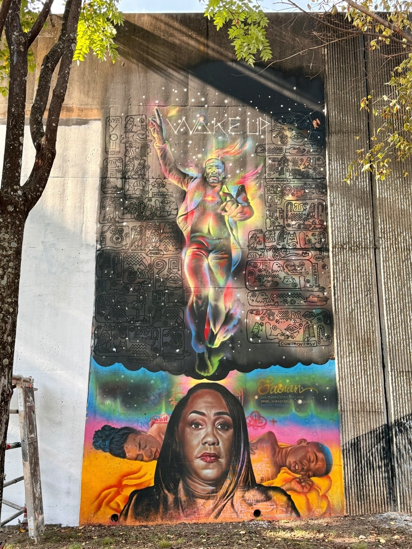 This image is a vibrant mural painted on a concrete wall. The artwork features a colorful central figure in a dynamic pose with the words “WAKE UP” above. Surrounding this figure are abstract and symbolic patterns.