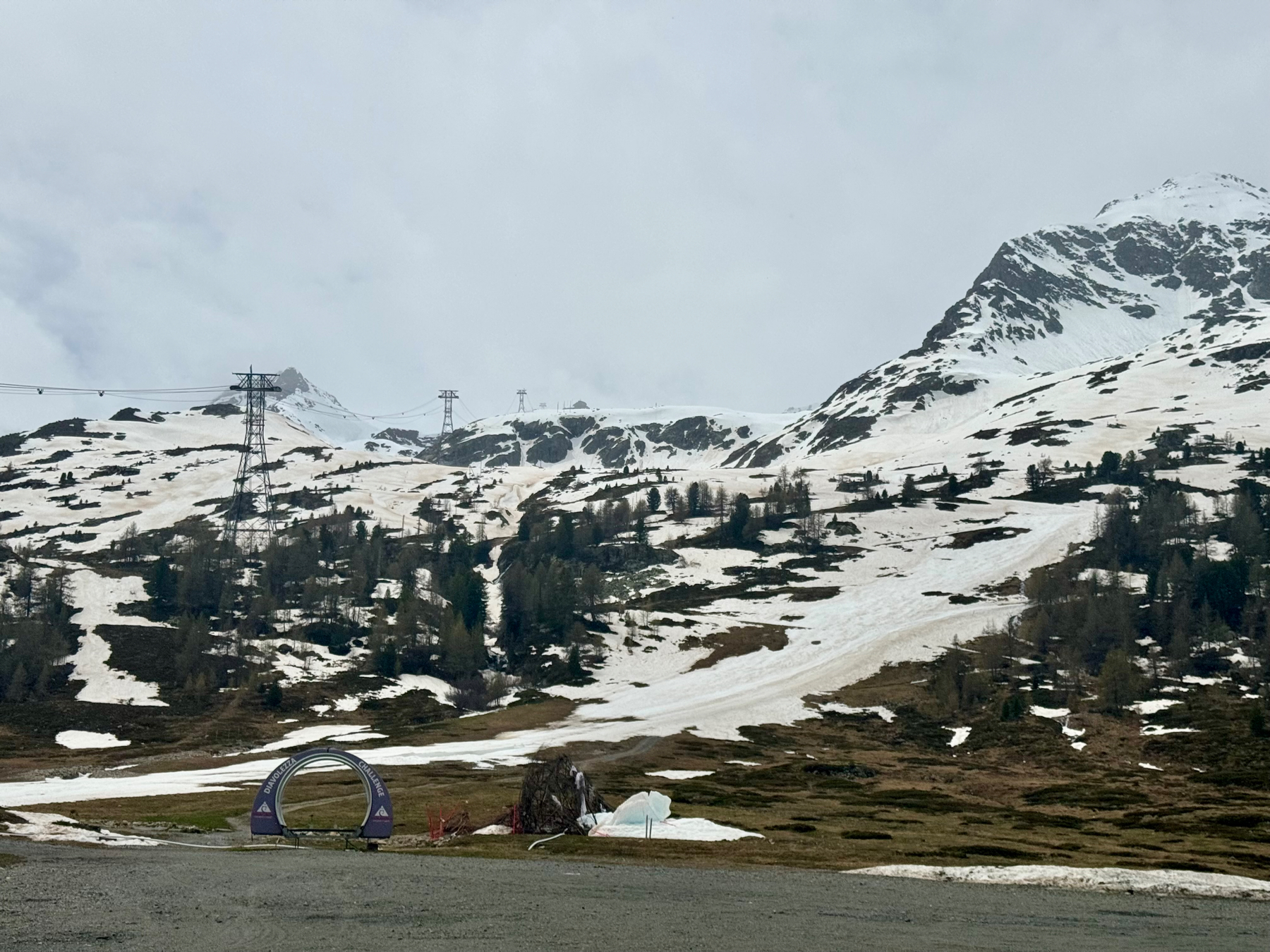 Snow-patched alpine landscape with a ski chairlift system and a partial view of a tunnel structure in the foreground. Cloudy skies suggest overcast weather.