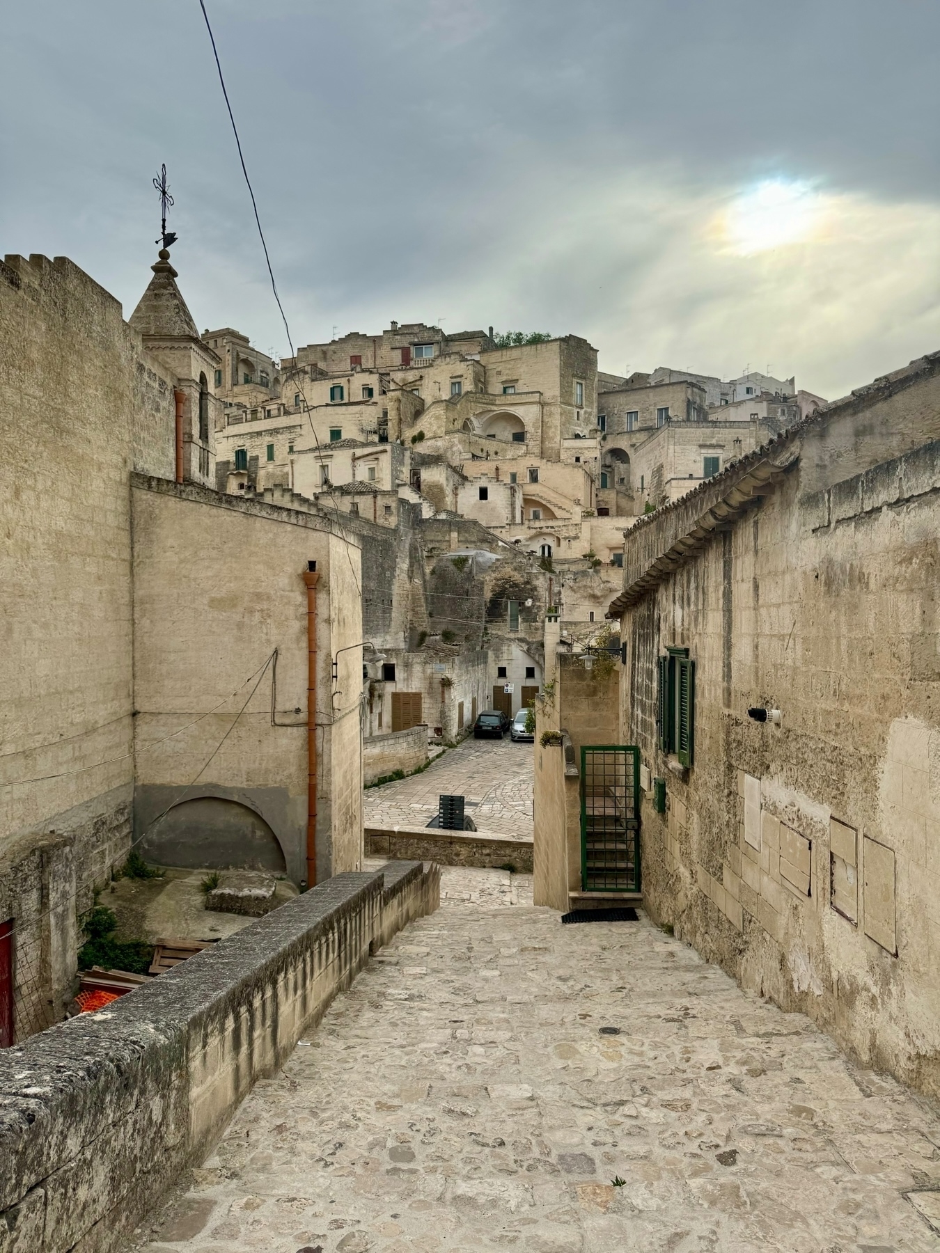 Historic stone buildings densely packed on a hillside under a cloudy sky. Stone-paved pathway descends through the middle, with a few parked cars visible at the bottom. The architecture features a mix of arches, stairs, and balconies with weathered textures.