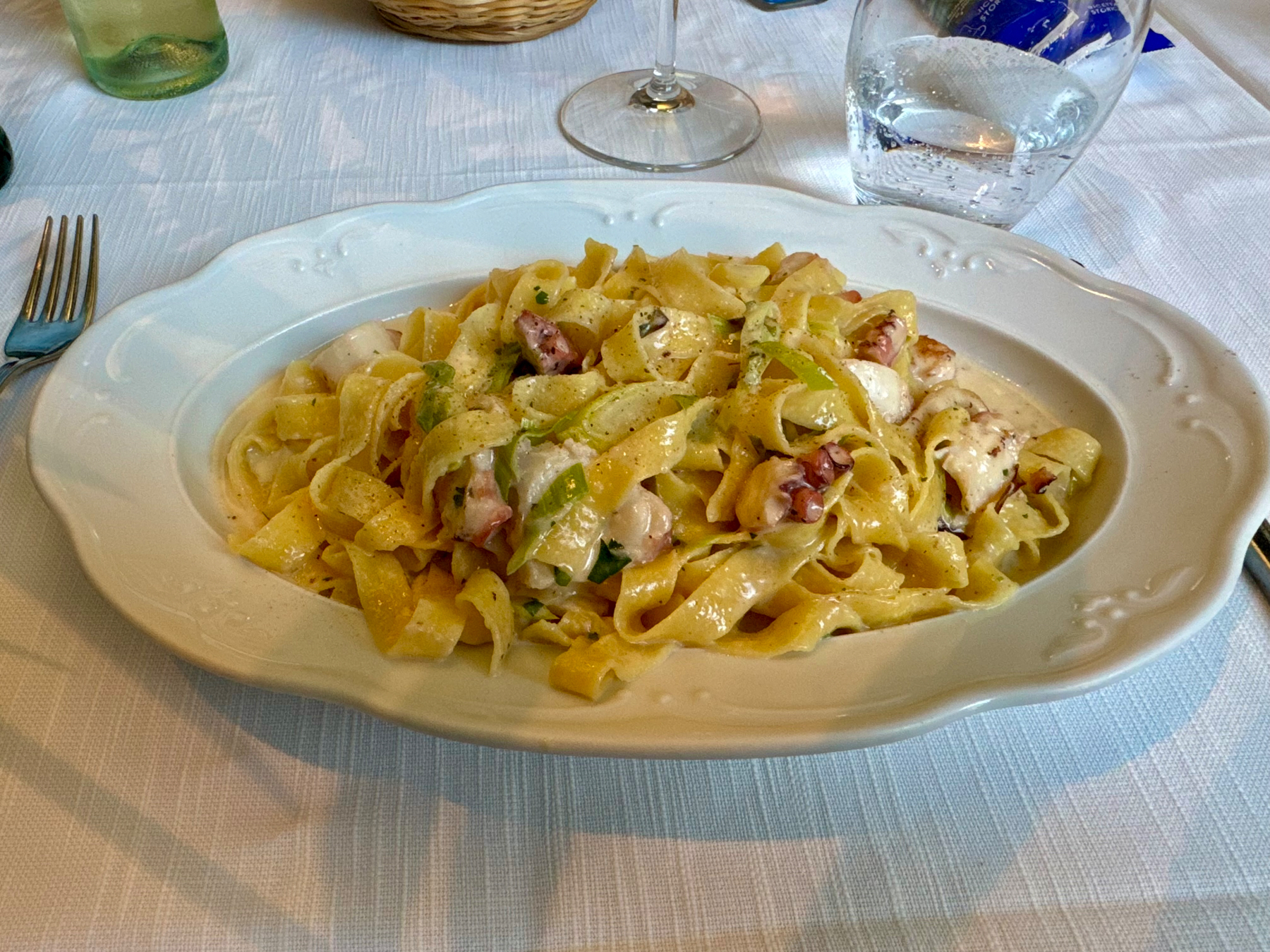 A white plate filled with creamy pasta dish, tagliatelle, garnished with octopus. The plate is on a white tablecloth with a fork, wine glass, and glass of water visible around it.