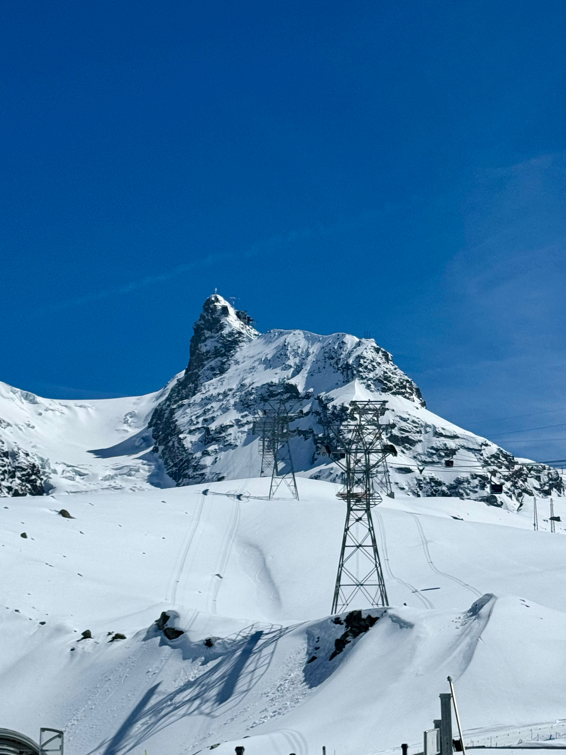Snow-covered mountain peak against a clear blue sky, with ski tracks, electricity pylons, and a partial view of a lift station at the base.