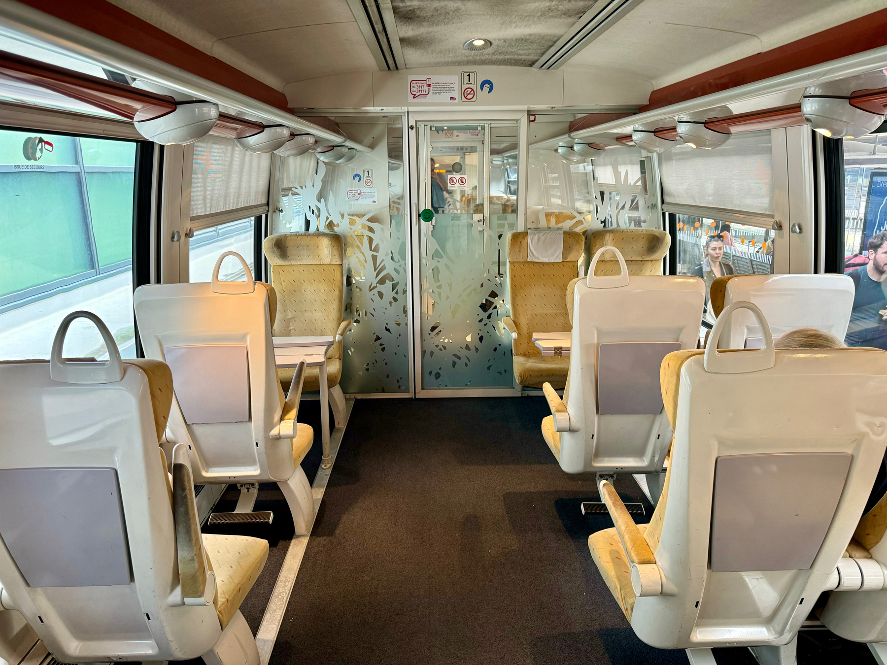 Interior of a train carriage with empty seats, patterned divider, overhead luggage compartments, and passengers seated further down.