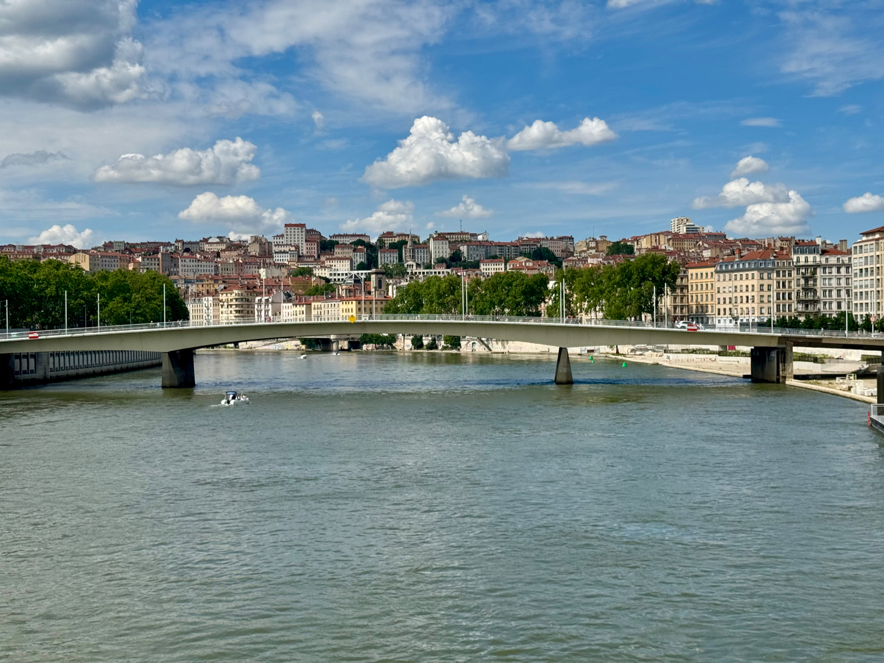 Lyon, France, showing the Sôane river with a motorboat passing under a concrete bridge. The bridge has streetlights and connects urban areas with historic buildings and dense greenery on the riverbanks. The sky is partly cloudy with blue patches.