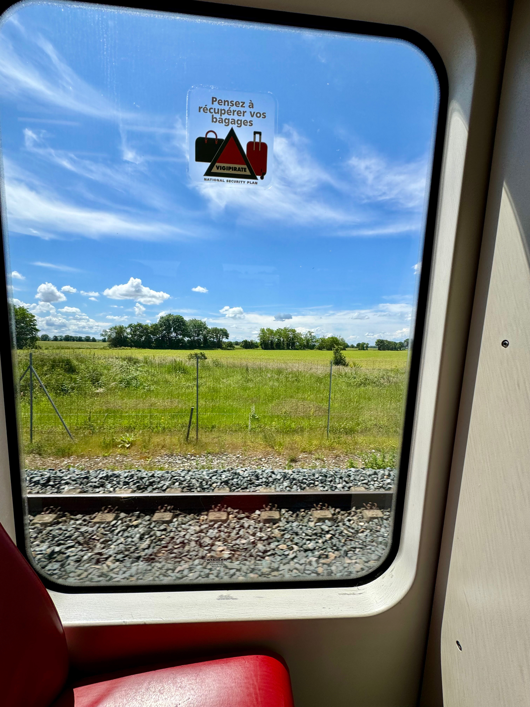 View from inside a train with a window sticker reminding passengers to collect their luggage, featuring the logo “Vigipirate,” the French national security alert system. Beyond the window are train tracks, green fields, and a blue sky with clouds.