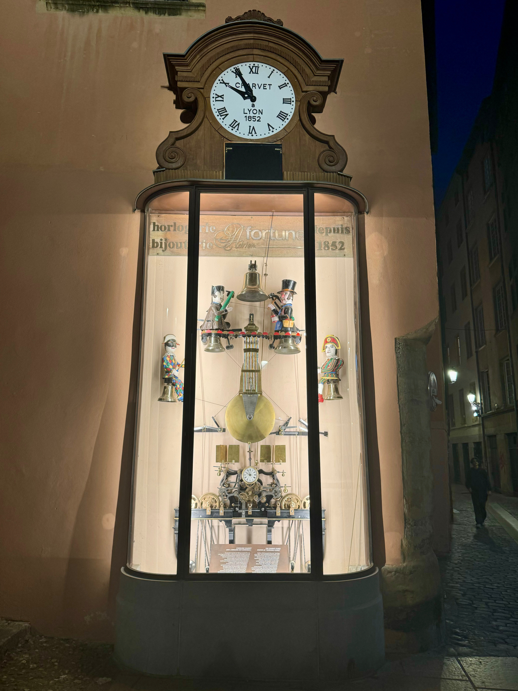 A clock shop window display at night featuring a large overhead wall clock with the brand “CRIVET” and “LYON 1852” and various clock mechanisms and artistic objects inside.